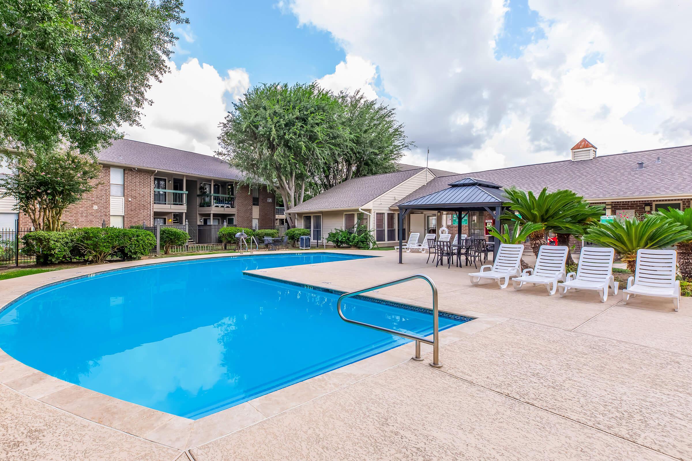 A clear blue swimming pool surrounded by lounge chairs and palm trees, with a gazebo in the background. Apartment buildings are visible to the left, and the sky is partly cloudy, creating a sunny and inviting atmosphere.