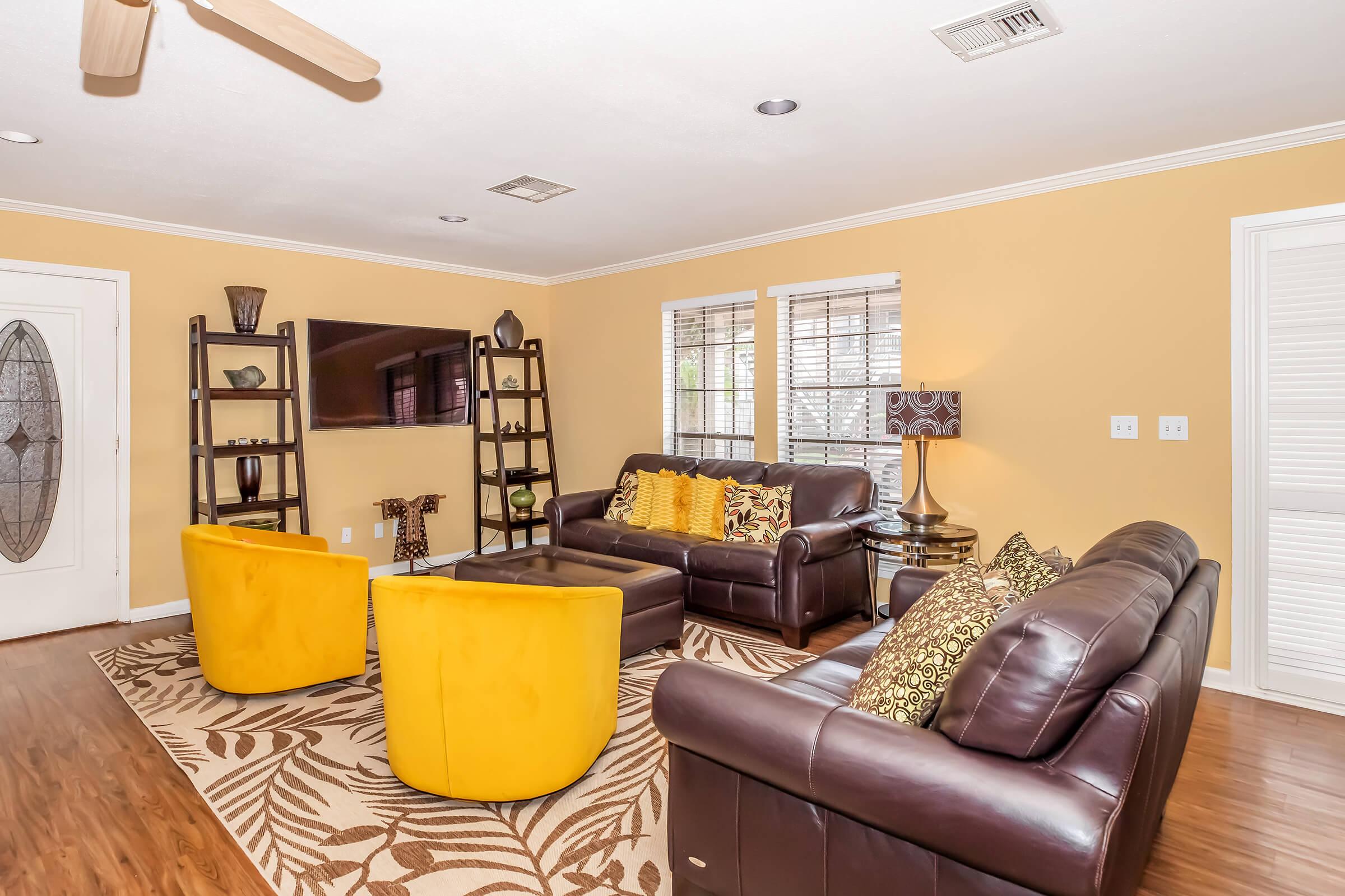 A cozy living room featuring a yellow and brown color scheme. Two bright yellow armchairs and a matching ottoman complement dark leather sofas. A modern bookshelf displays decorative items, while a large TV is mounted on the wall. Windows with blinds allow natural light, and a patterned area rug adds warmth to the space.