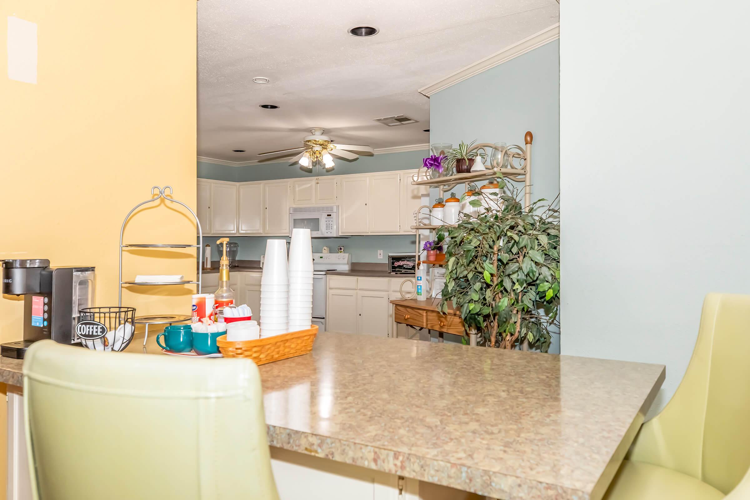 A bright kitchen area featuring a countertop with coffee cups and condiments, a small basket, and a coffee station. The background shows white cabinets, a ceiling fan, and a plant beside a wooden shelf filled with kitchen items. The walls are painted in soft pastel colors, creating a welcoming atmosphere.