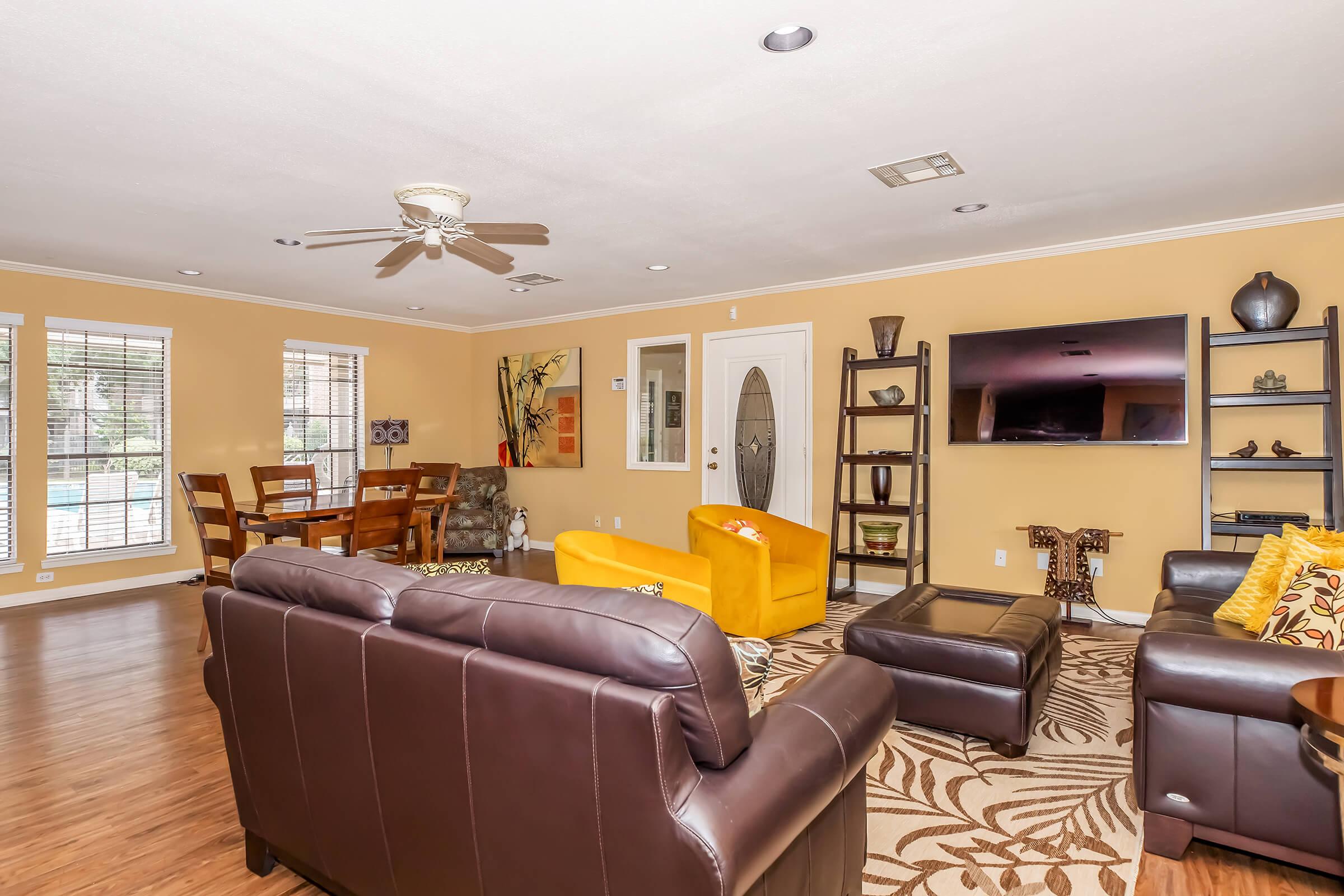 A spacious living room featuring a brown leather sofa, an orange armchair, and a coffee table. There is a TV mounted on the wall and decorative shelves. Large windows allow natural light, and a dining area with a wooden table is visible in the background. The walls are painted a warm tan color.