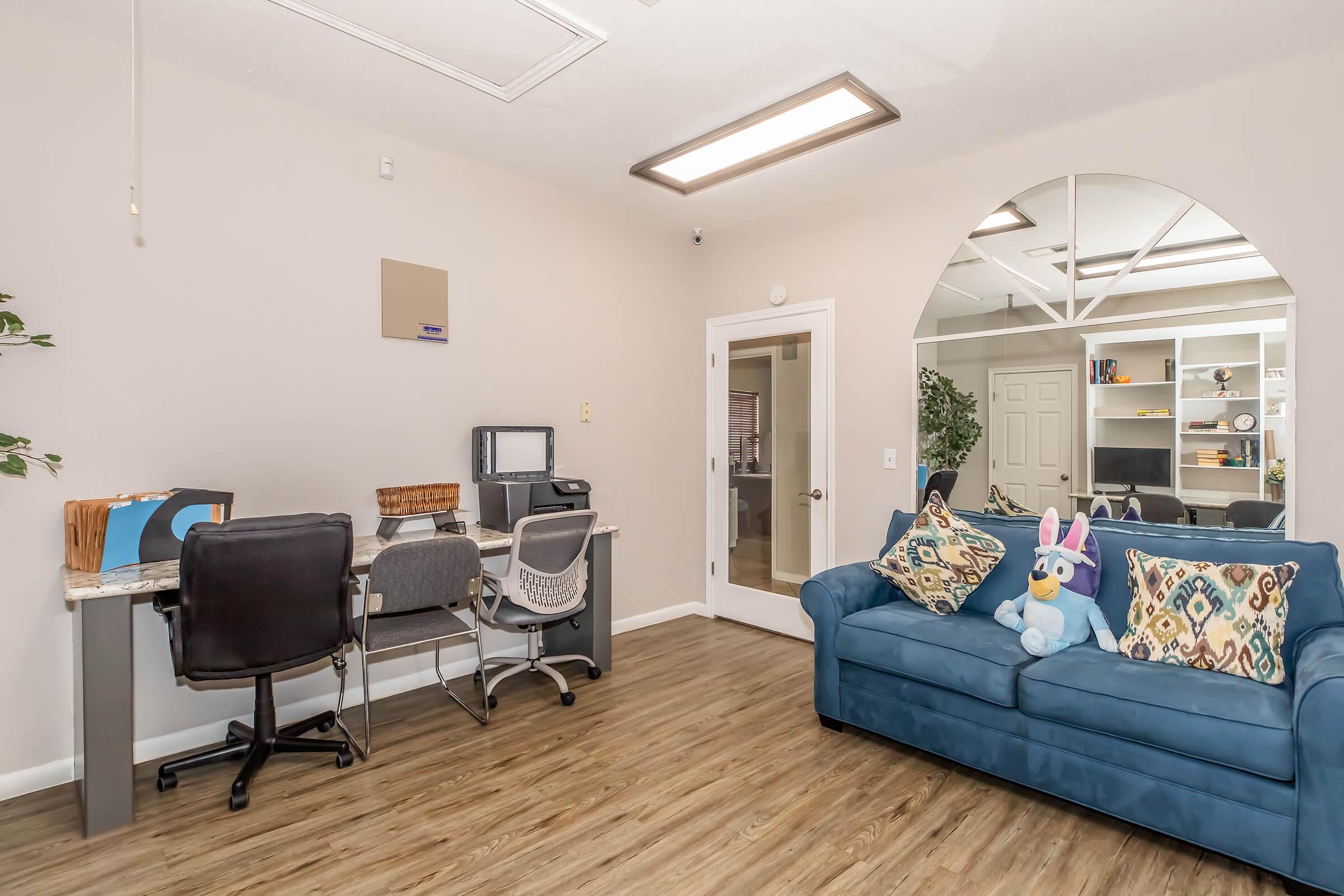 A cozy office space featuring a blue couch with colorful pillows, a small desk with a computer and chair, and a decorative window archway. A playful stuffed bunny is placed on the couch, and there are plants and a bookshelf in the background, creating a welcoming and comfortable work environment.