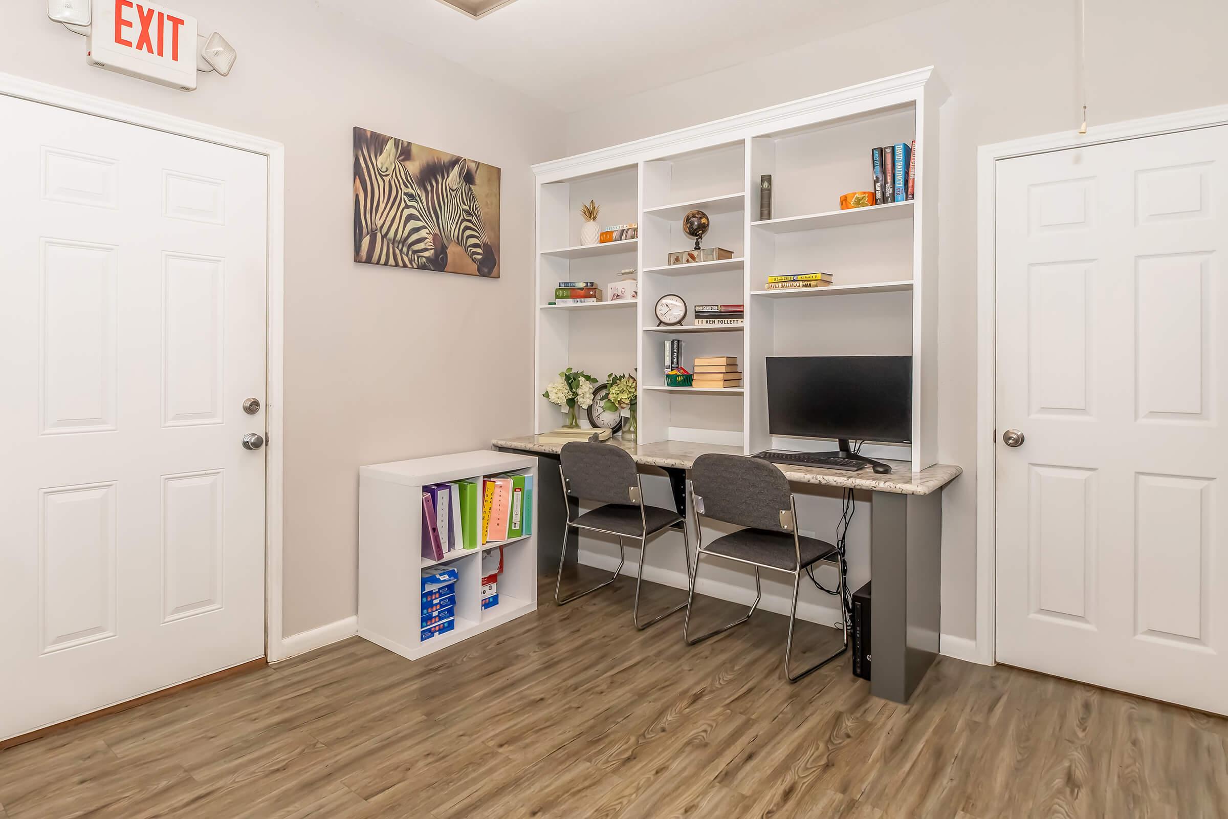 A small office space featuring a desk with two chairs, a computer monitor, and shelves filled with books and decorative items. The walls are light-colored, and there’s a zebra-themed painting above the desk. Two doors are visible, one labeled "EXIT," and the floor has a wood-like finish.