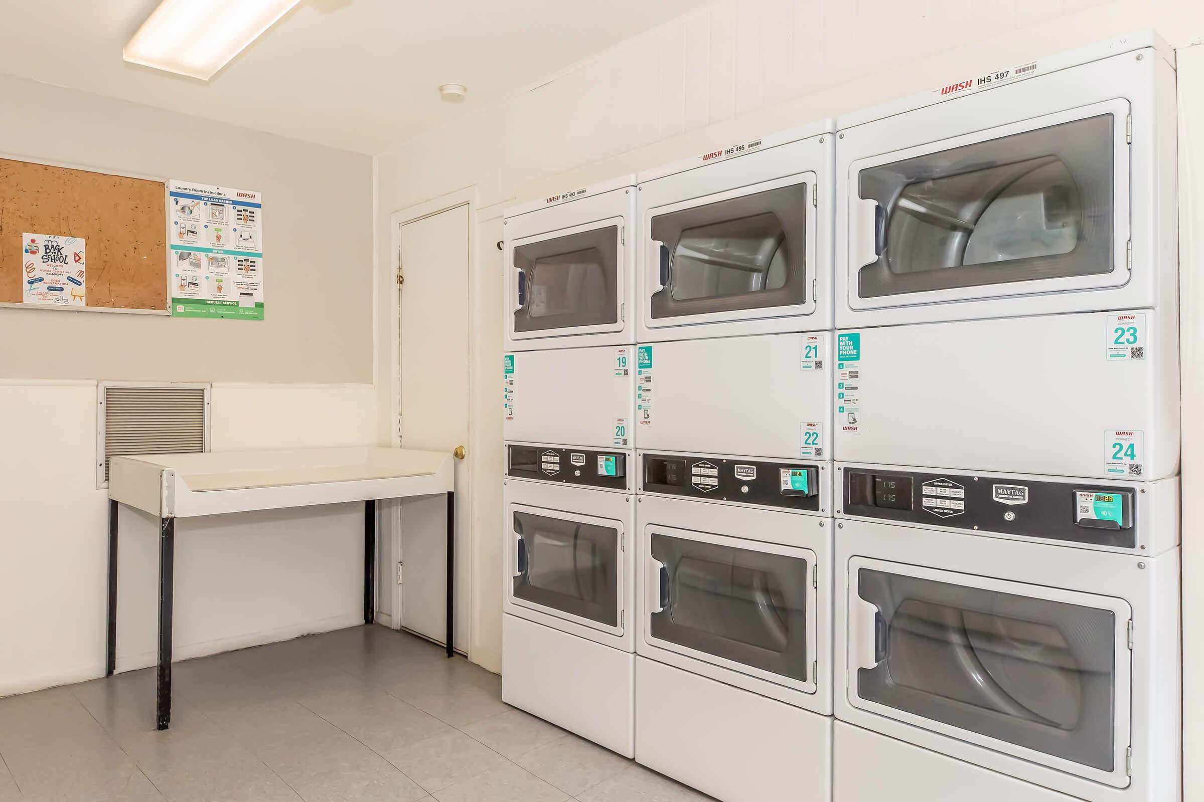 A clean laundry room featuring stacked washing machines and dryers against a wall. There is a folding table to the left and a bulletin board on the wall, displaying laundry-related information and notices. The space is well-lit and organized for convenience.