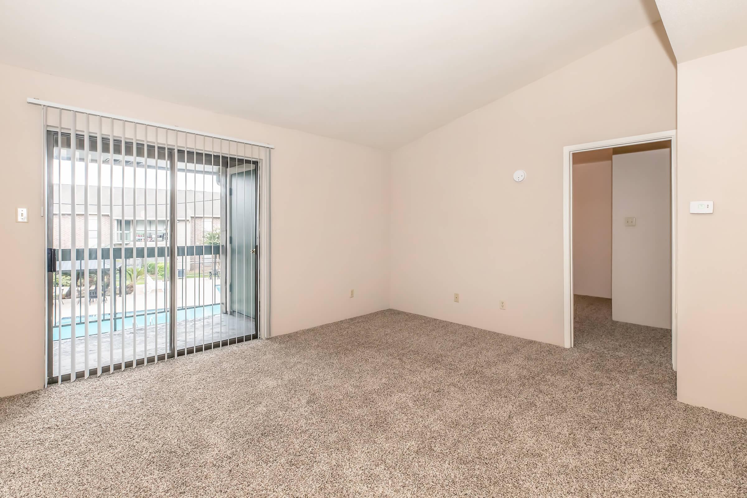 Empty living room with beige walls and plush carpet. A sliding glass door with vertical blinds leads to an outdoor area. There is a doorway on the right leading to another room. Natural light enters through the glass door, illuminating the space.