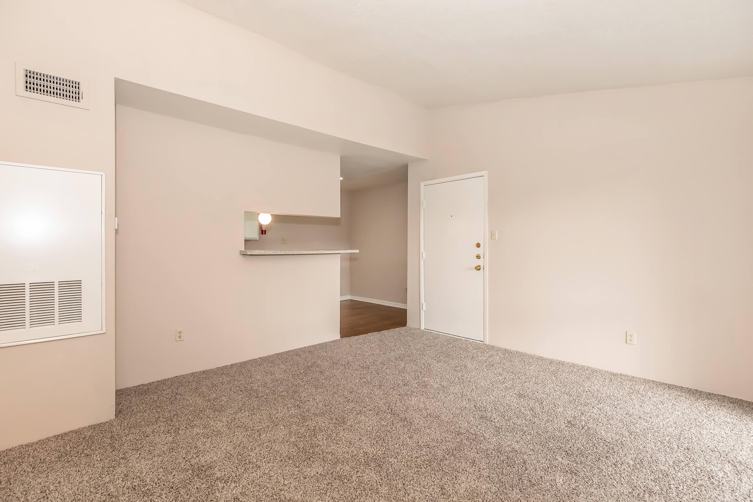 A spacious, light-colored room with beige walls and a soft carpet. The area features an open layout leading to a doorway on the right and a small pass-through to a kitchen area in the background. The door is white with a round doorknob, and there is a wall vent to the left.