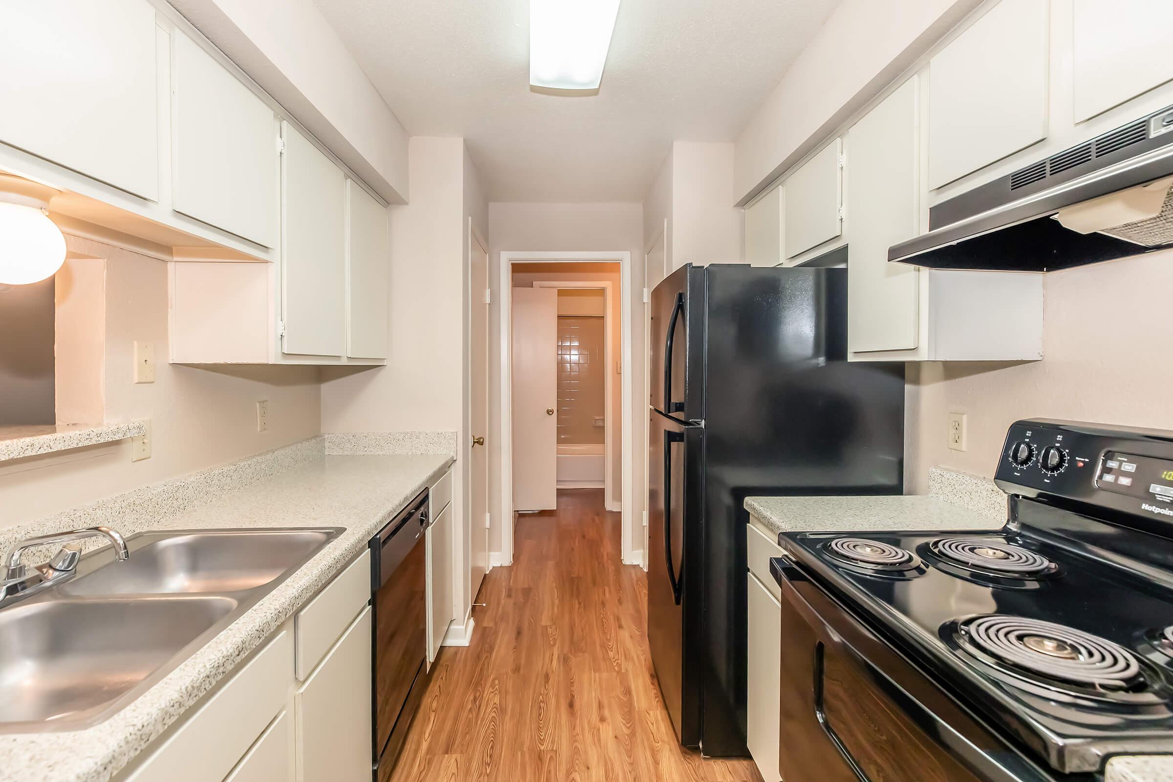 A modern kitchen featuring white cabinetry, a double sink, black appliances including a refrigerator and stove, and granite countertops. The space has wood laminate flooring and leads to a hallway with a door visible in the background, indicating access to other areas of the home.