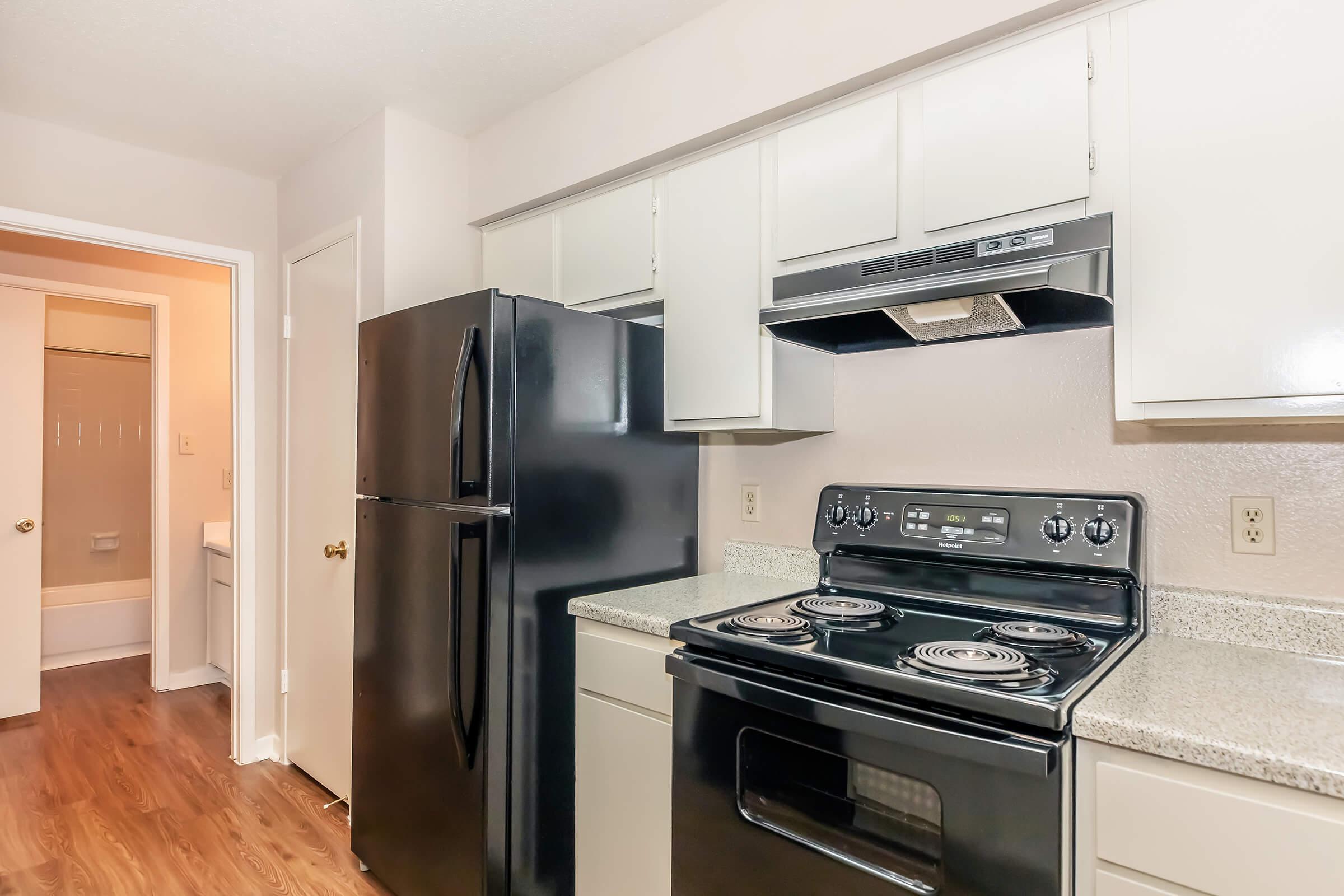 Modern kitchen featuring black appliances including a refrigerator and stove, granite countertops, and light-colored cabinetry. A doorway in the background leads to a bathroom, and the flooring is a warm, wood-like laminate.