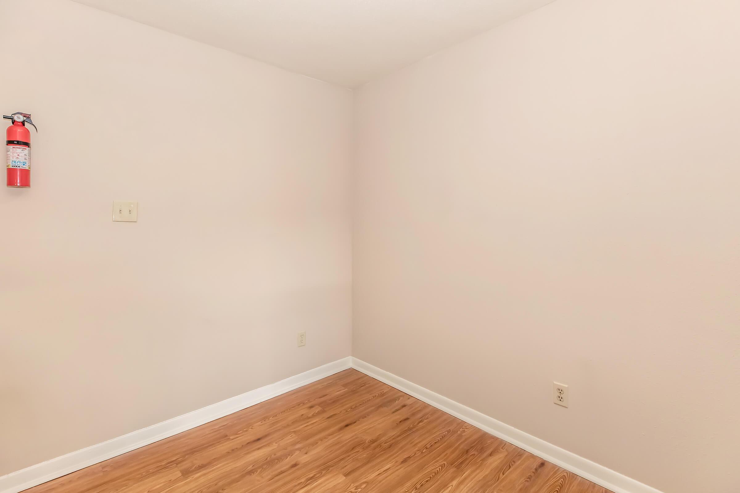 Empty room with light pink walls and a wooden floor. A fire extinguisher is mounted on the wall to the left. No furniture or decorations are present, creating a minimalistic and open space.