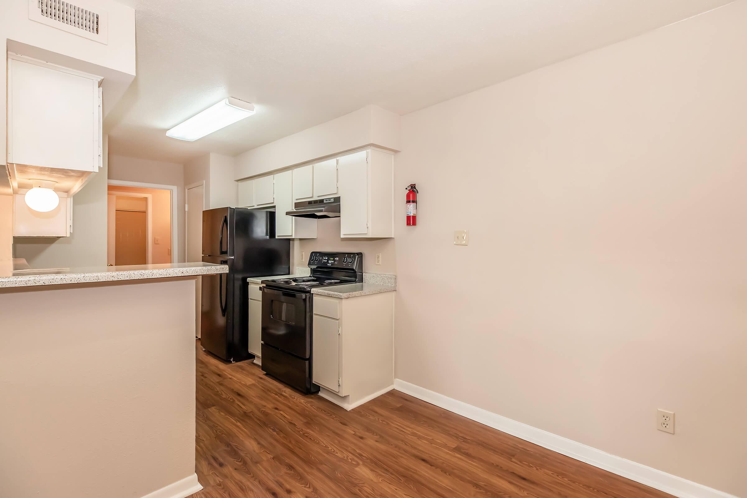 A clean, modern kitchen with white cabinets, black appliances including a refrigerator and a stove, and laminate flooring. The kitchen has a small bar area opening to an adjacent room, with neutral-colored walls and ample light from a ceiling fixture. A fire extinguisher is mounted on the wall.