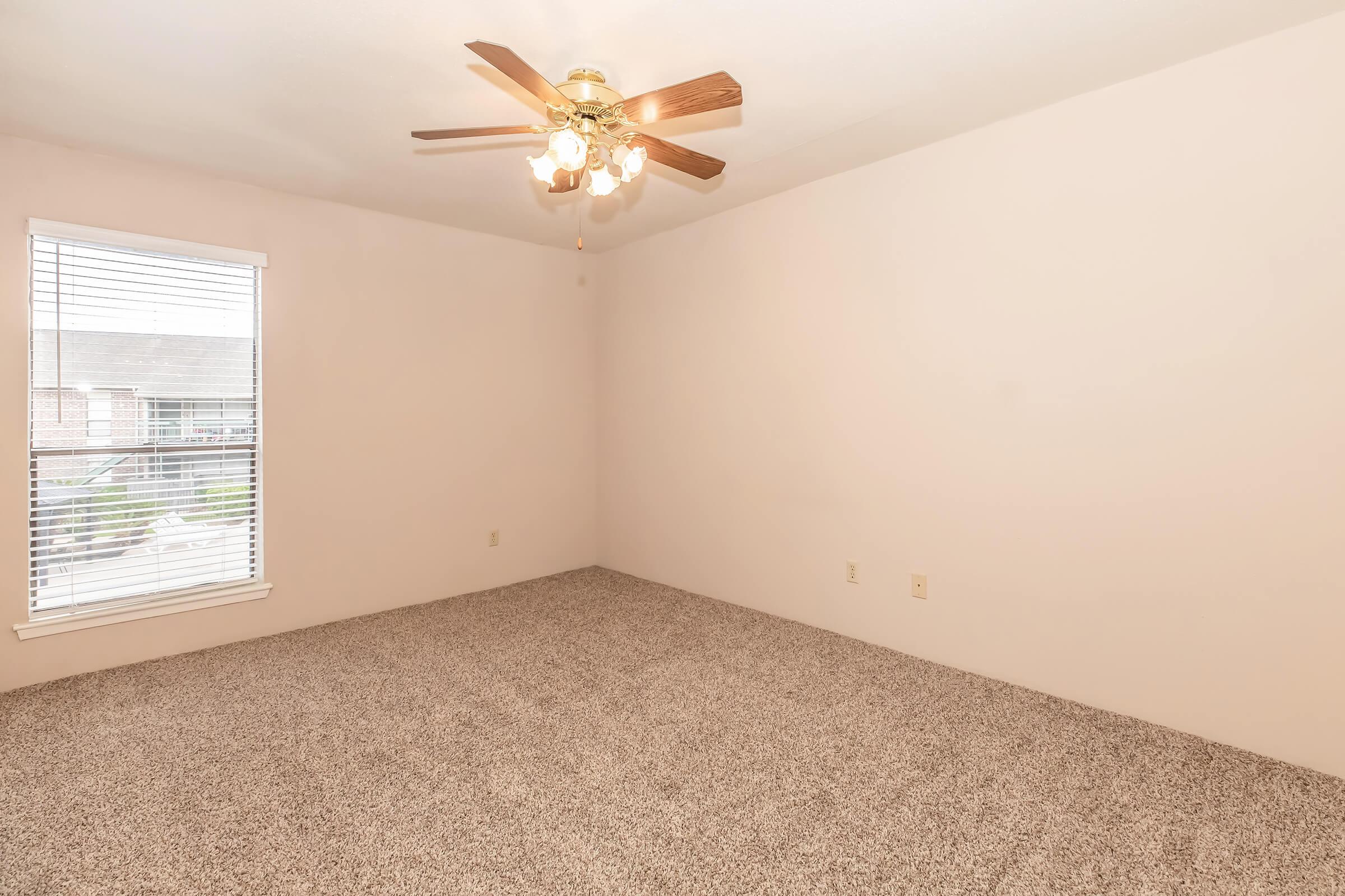 Empty room featuring light beige walls and a ceiling fan with wooden blades. The floor is covered in gray carpeting, and there is a window with blinds allowing natural light. The space appears bright and airy, with no furniture or decorations.