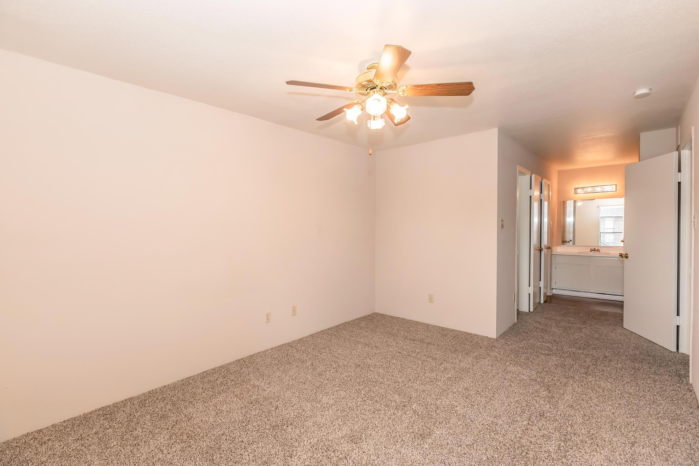 A spacious, empty room featuring light-colored walls and a ceiling fan with four lights. The flooring is covered with gray carpet. A doorway leads to a bathroom area with a mirror and vanity, visible in the background. The overall ambiance is bright and inviting, suitable for various setups.