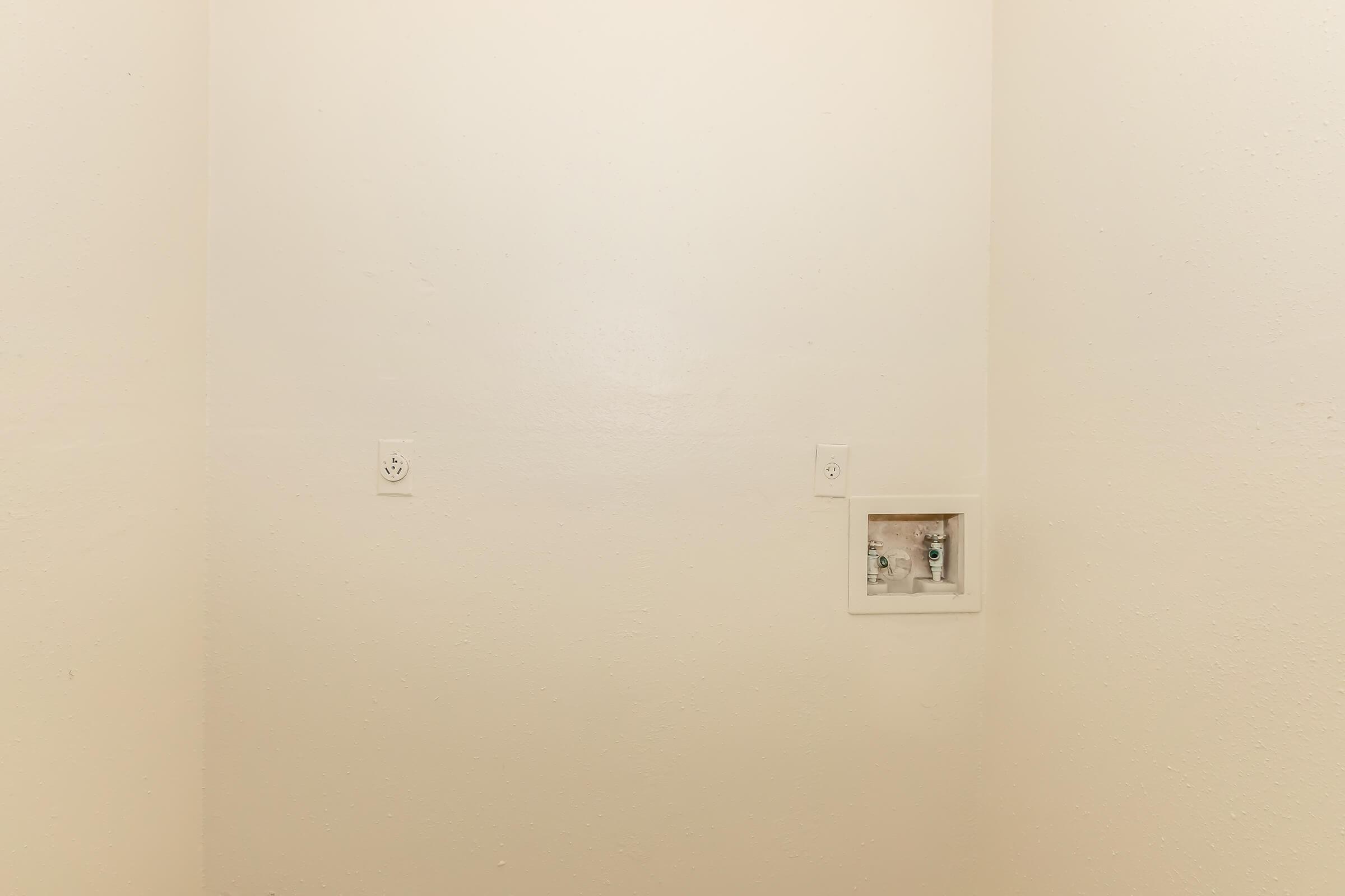 A plain white wall with two electrical outlets and a small electrical box visible on the lower right side. The surface is smooth with minimal texture, creating a simple and unadorned appearance.