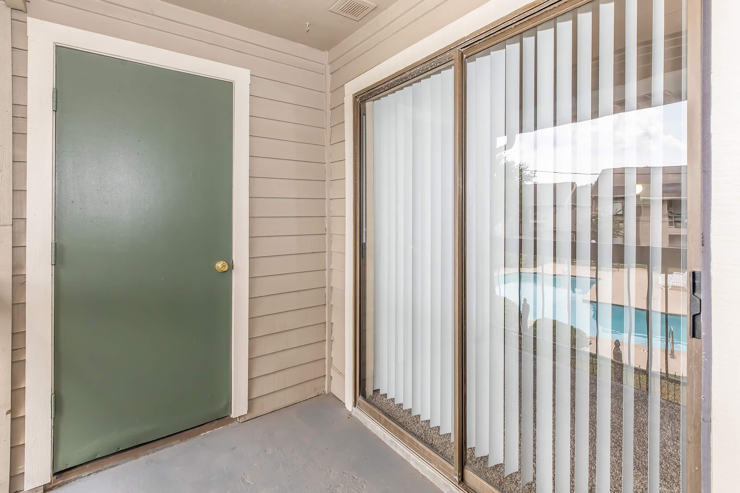 A cozy entrance with a green door, leading to a patio with sliding glass doors. The glass doors feature vertical blinds that are partially open, revealing a glimpse of a pool area in the background. The walls are light-colored wood paneling, creating a warm and inviting atmosphere.