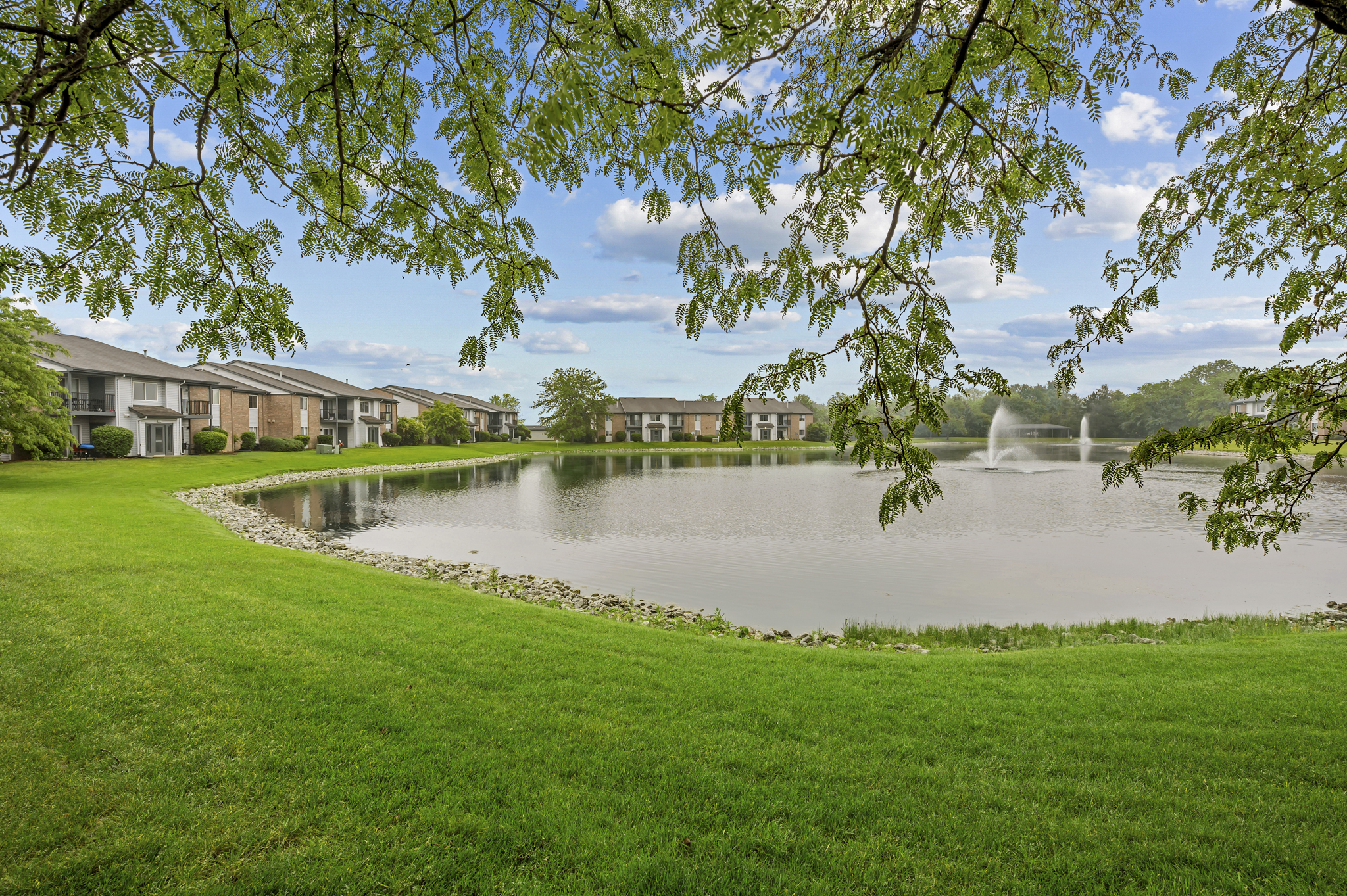 A serene lake surrounded by lush green grass and trees, with a fountain in the water. In the background, there are modern apartment buildings reflecting a peaceful residential setting, under a bright blue sky with scattered clouds.