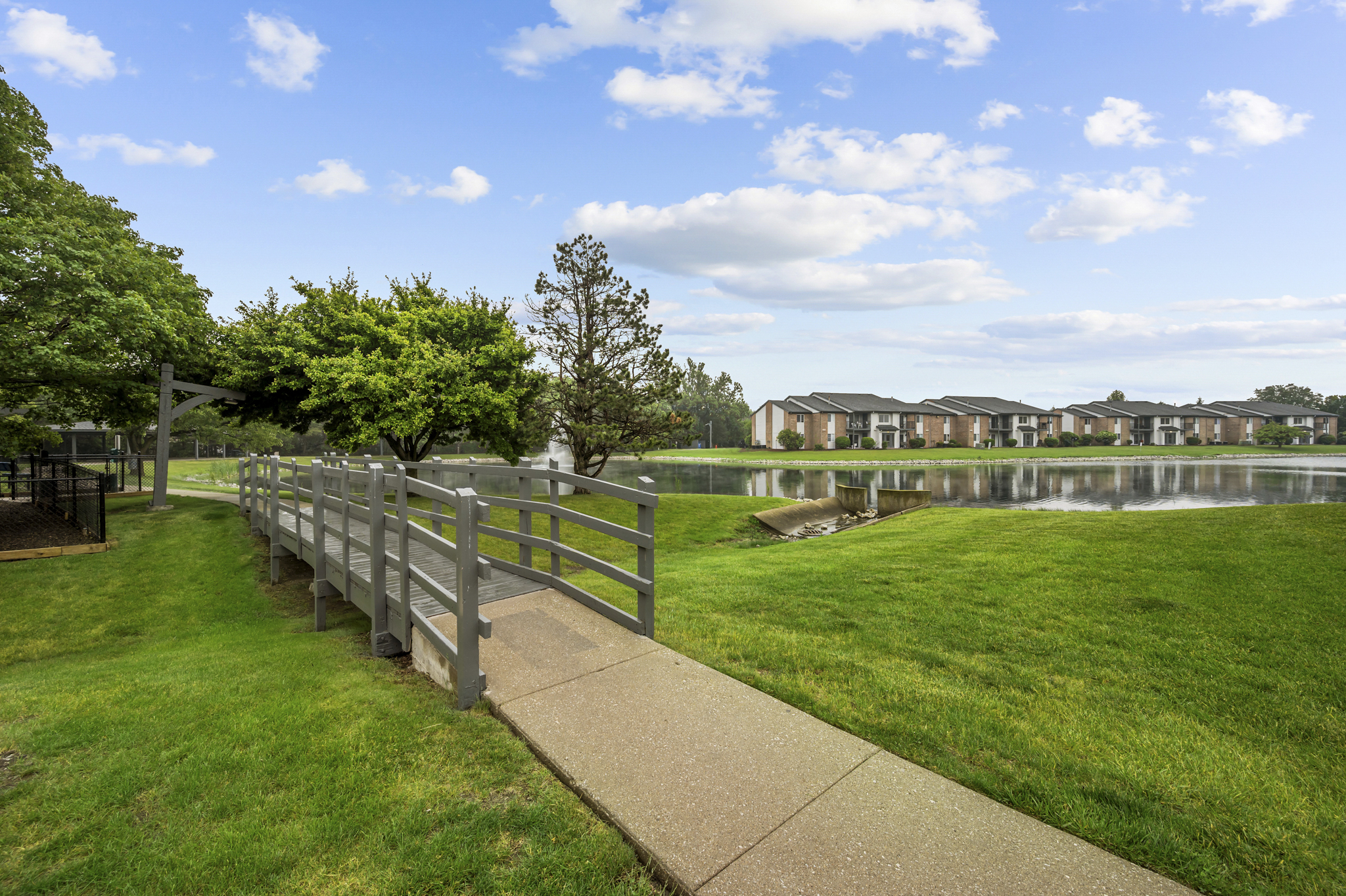 A peaceful landscape featuring a wooden pathway leading over a pond. Lush green grass surrounds the area, with trees and shrubbery on one side and modern apartment buildings reflected in the calm water. The sky is partly cloudy, enhancing the serene atmosphere.