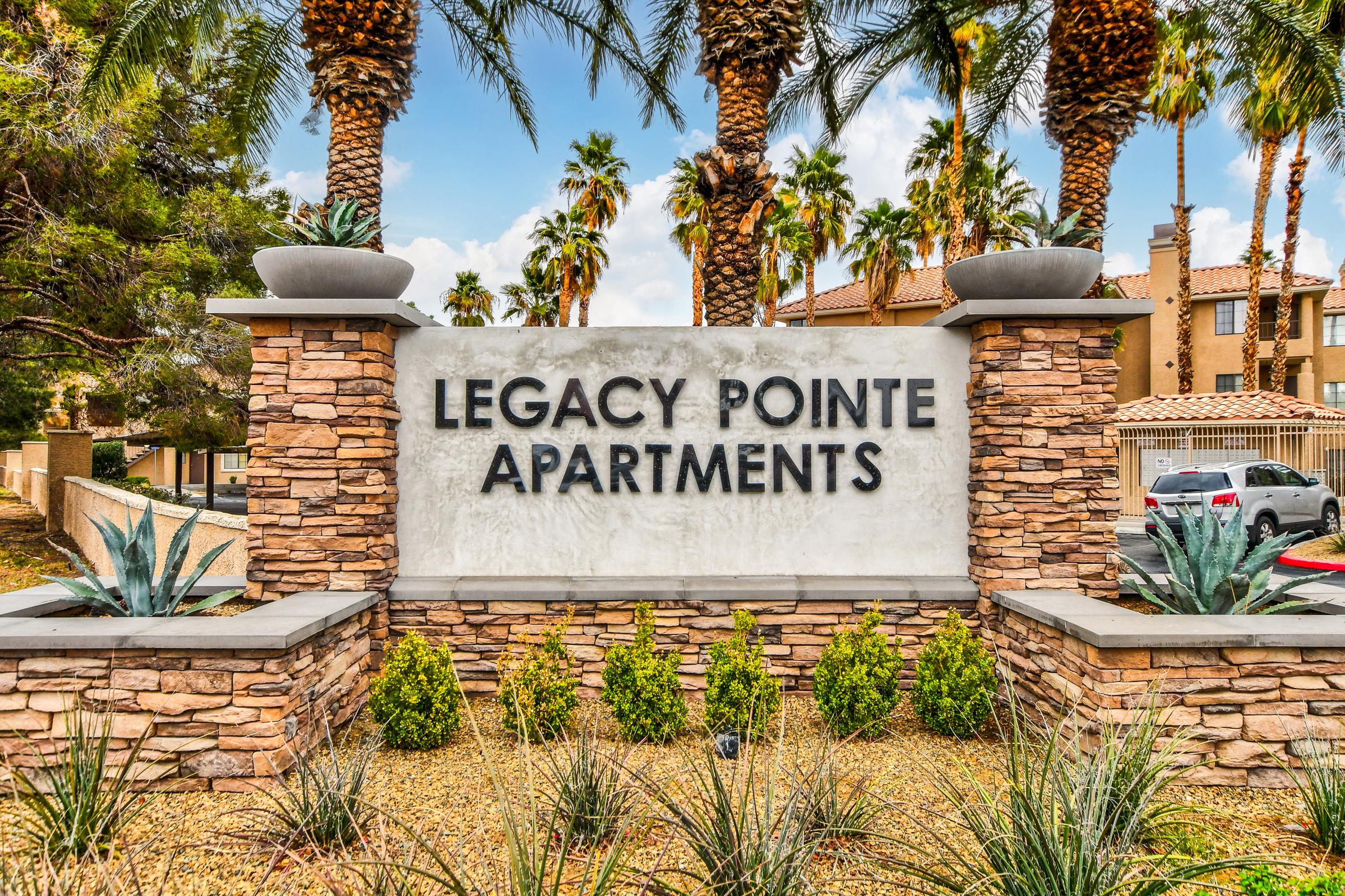 Sign for Legacy Pointe Apartments featuring stylized lettering. Surrounded by palm trees and desert landscaping, the sign is made of stone and concrete, indicating the entrance to the apartment complex.