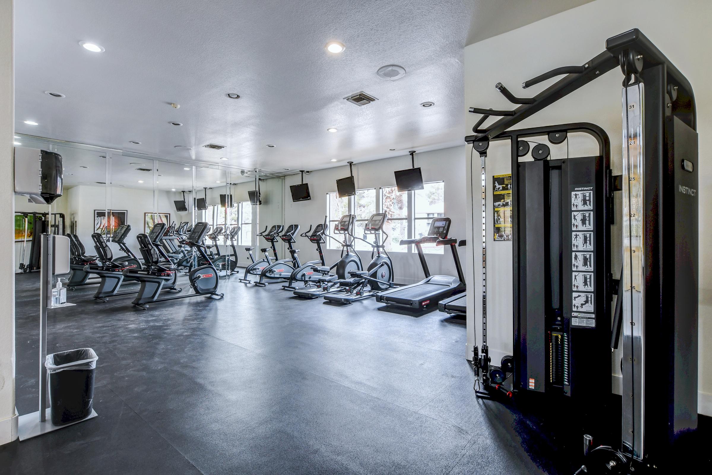 A modern gym interior featuring several cardio machines, including treadmills and stationary bikes, along with a multi-gym strength training machine. The space is well-lit with mirrors on one wall and multiple television screens mounted above the equipment. The flooring is black and the atmosphere looks inviting for workouts.