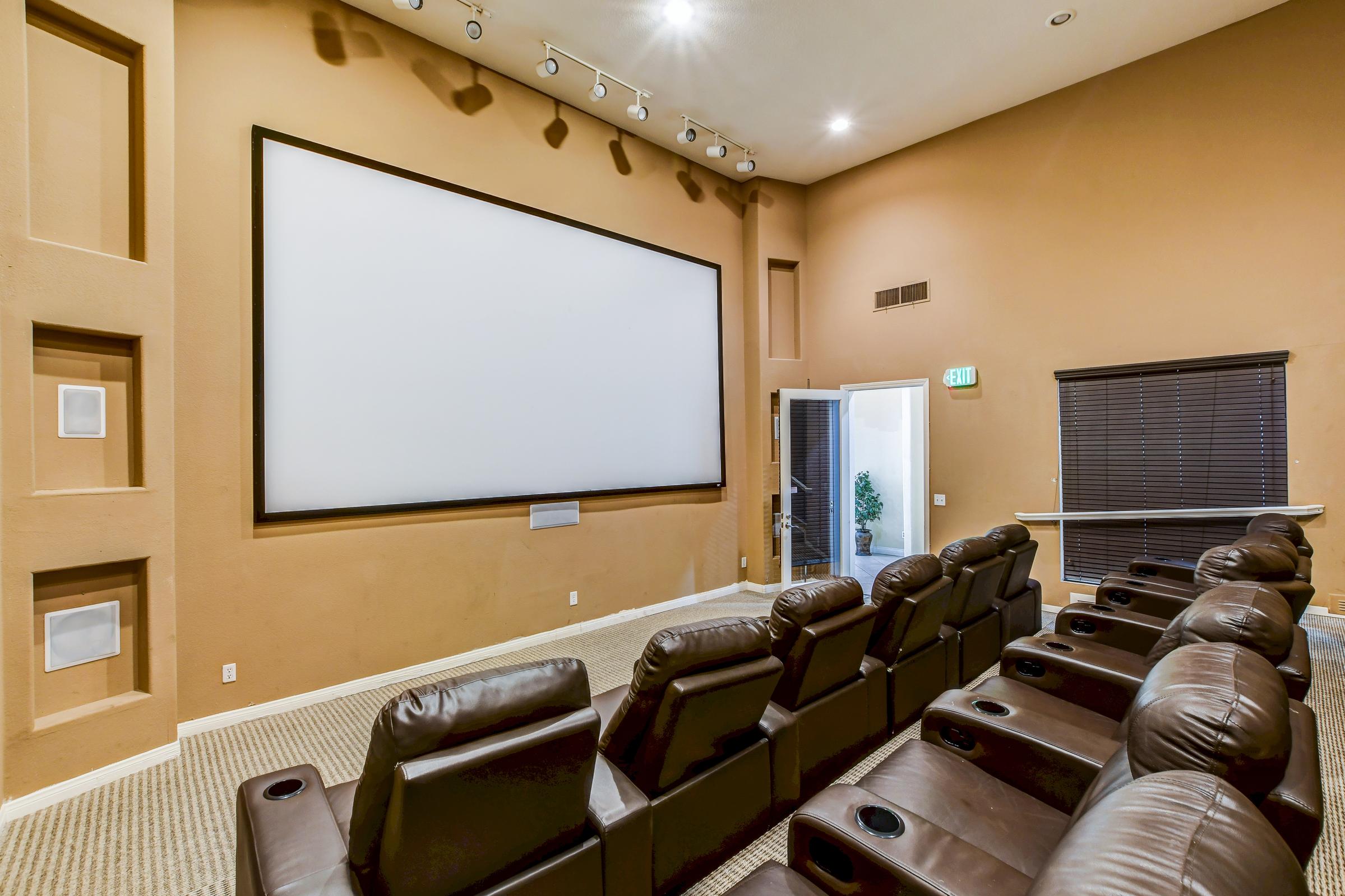 Home theater room featuring a large blank projection screen, plush leather reclining seats with cup holders, and warm-colored walls. Ambient lighting and soundproofing elements are visible, creating a cozy cinematic atmosphere. A doorway in the background leads to another space.
