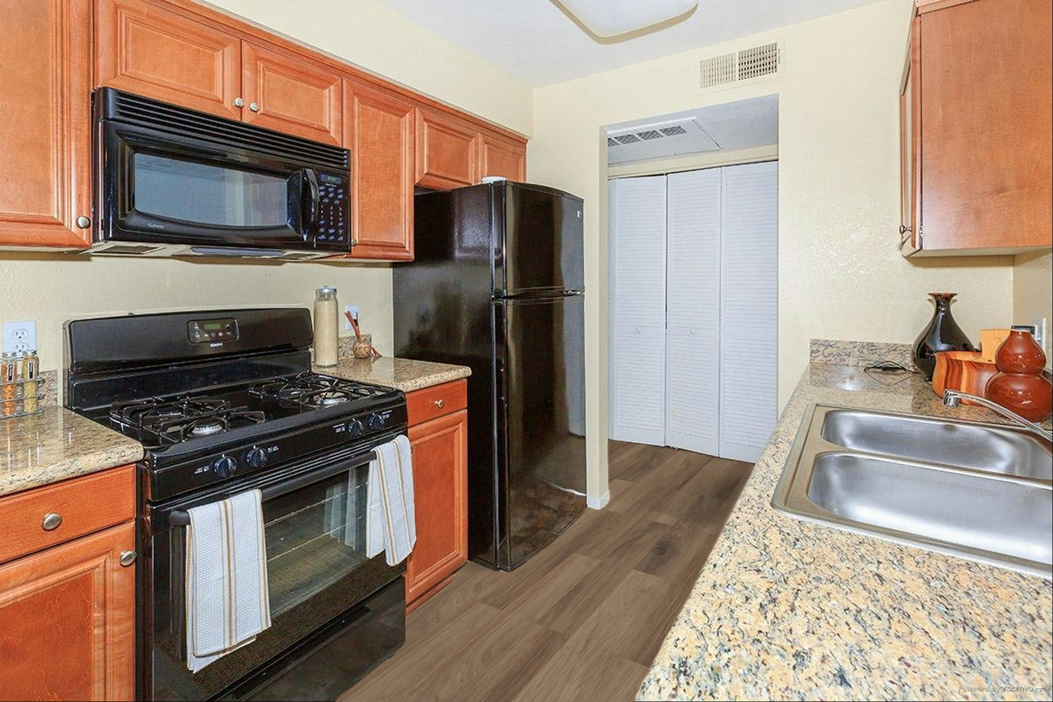 A modern kitchen featuring wooden cabinetry, a black microwave and refrigerator, and a gas stove. The countertops are granite with a brown and beige pattern. There are two sinks and a light-colored wall behind the appliances. A small hallway leads to a closet with double doors in the background.