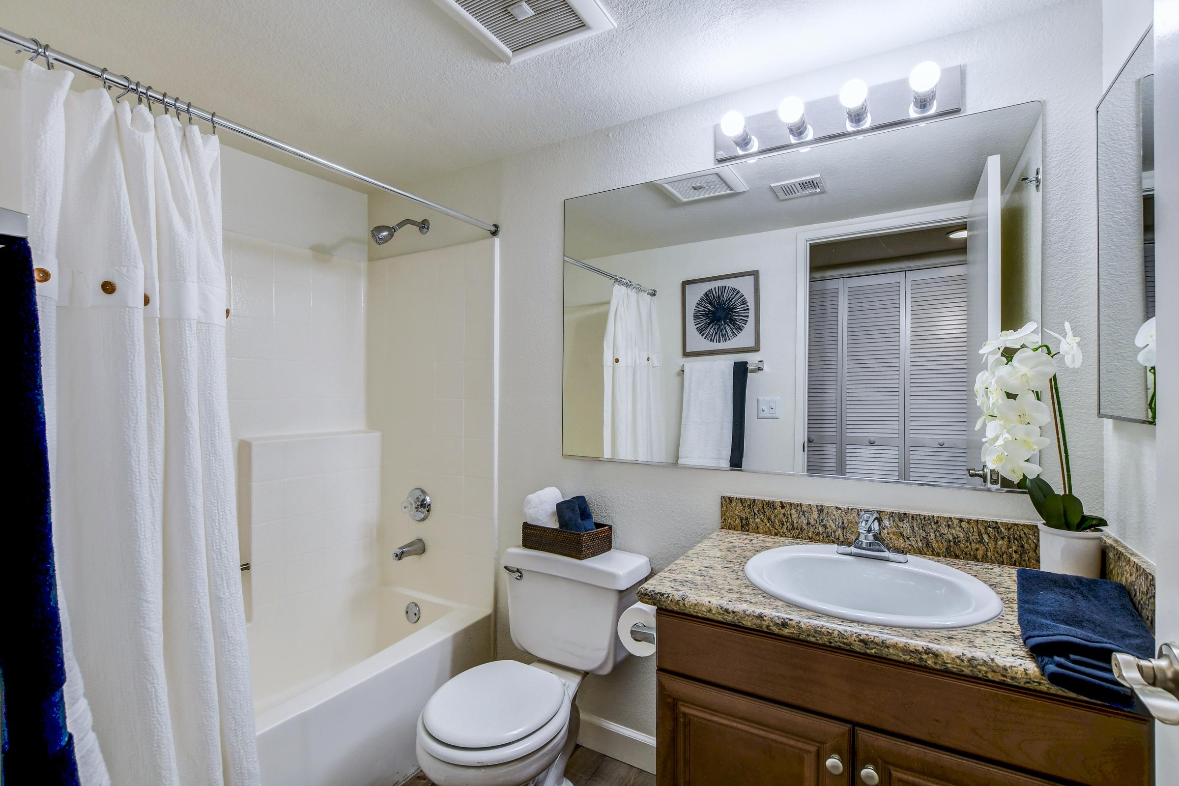 A modern bathroom featuring a white bathtub with a shower curtain, a granite countertop with a sink, and a toilet. The mirror above the sink reflects the room, which includes a decorative artwork on the wall and a window with blinds. White towels are neatly arranged on the counter.