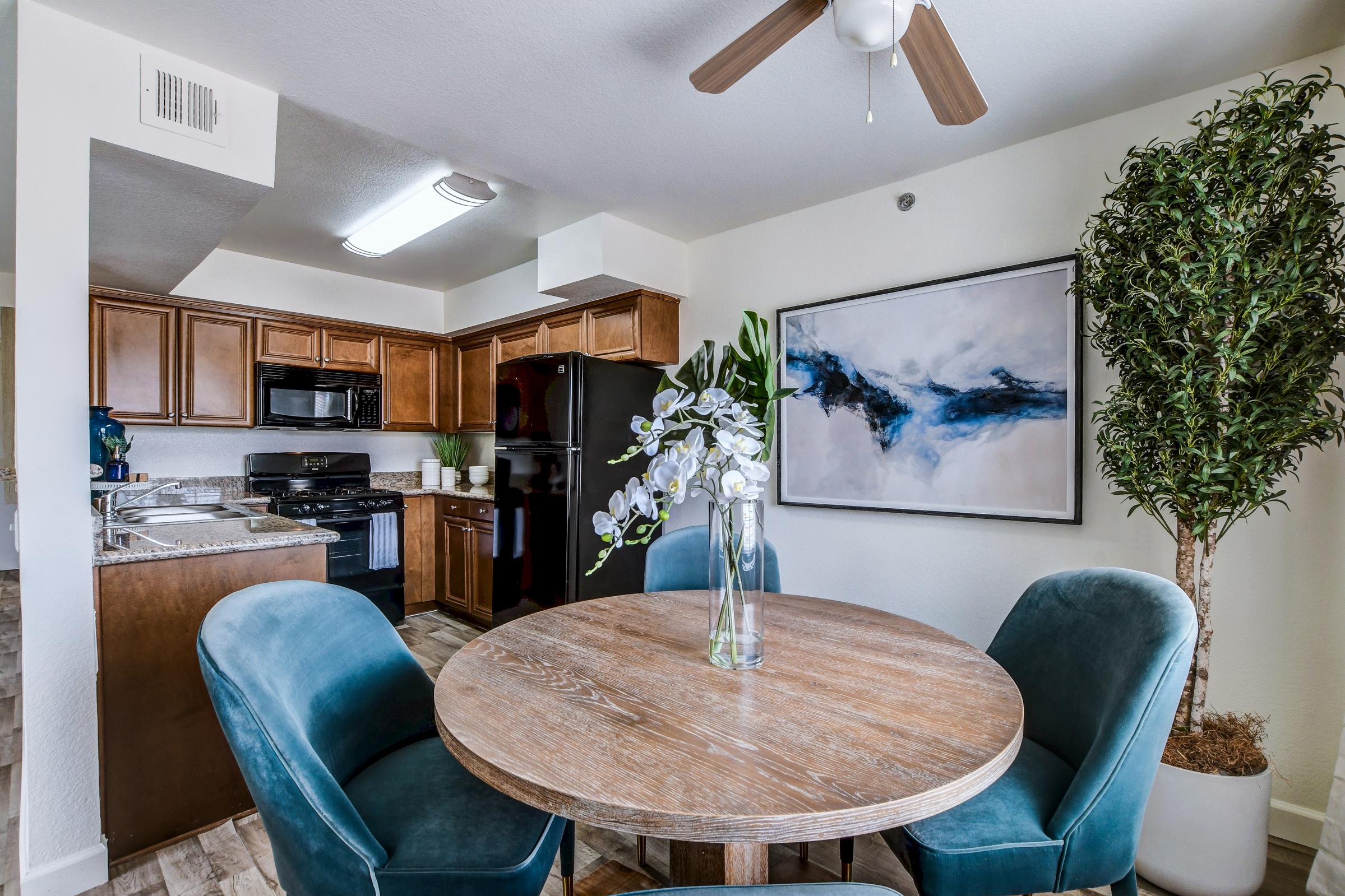 A modern kitchen with wooden cabinets and stainless steel appliances, featuring a round wooden dining table surrounded by teal upholstered chairs. A large abstract painting is on the wall, and a potted plant adds a touch of greenery to the space. Natural light brightens the room through a window.