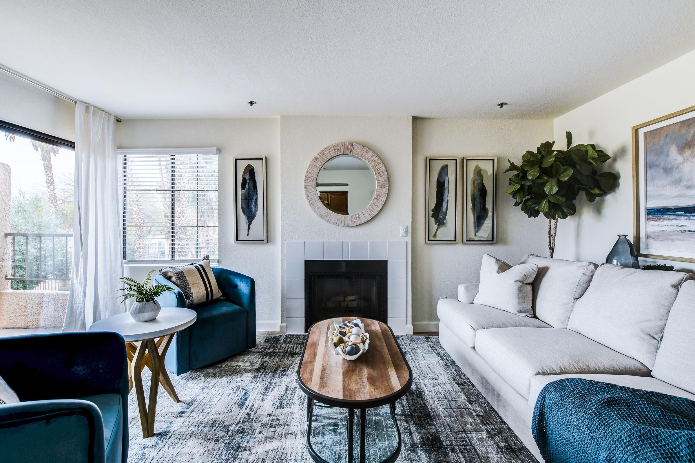A modern living room featuring a white sofa, blue accent chairs, and a wooden coffee table. The space includes a fireplace with a circular mirror above it, framed artwork on the walls, and decorative plants. Natural light streams in through a window, enhancing the cozy and inviting atmosphere.