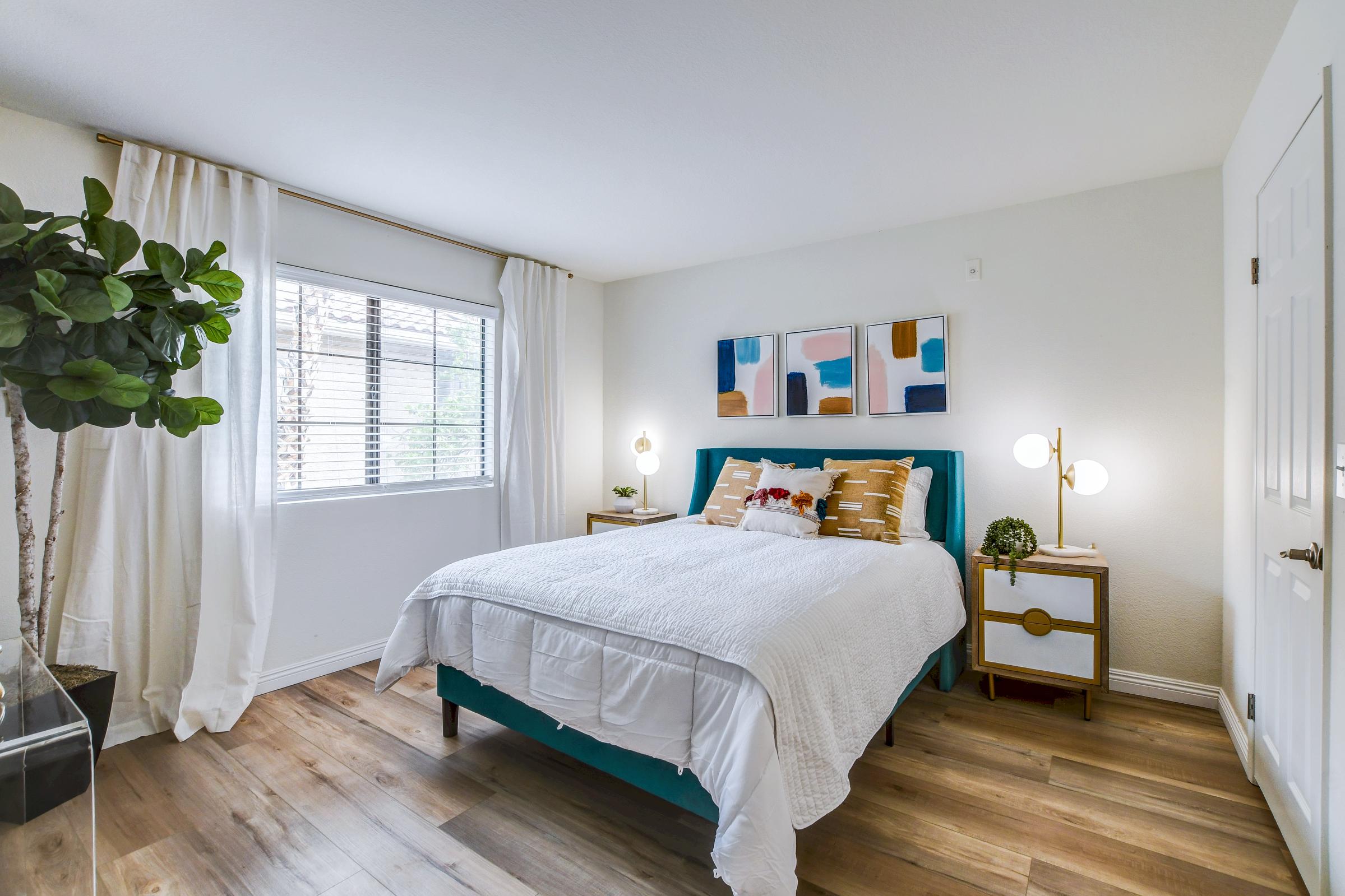 A cozy bedroom featuring a neatly made bed with white bedding and decorative pillows. There's a pair of stylish nightstands with decorative lamps on each side. A large window with sheer curtains lets in natural light, and a potted plant adds a touch of greenery to the space. The walls are adorned with abstract artwork.
