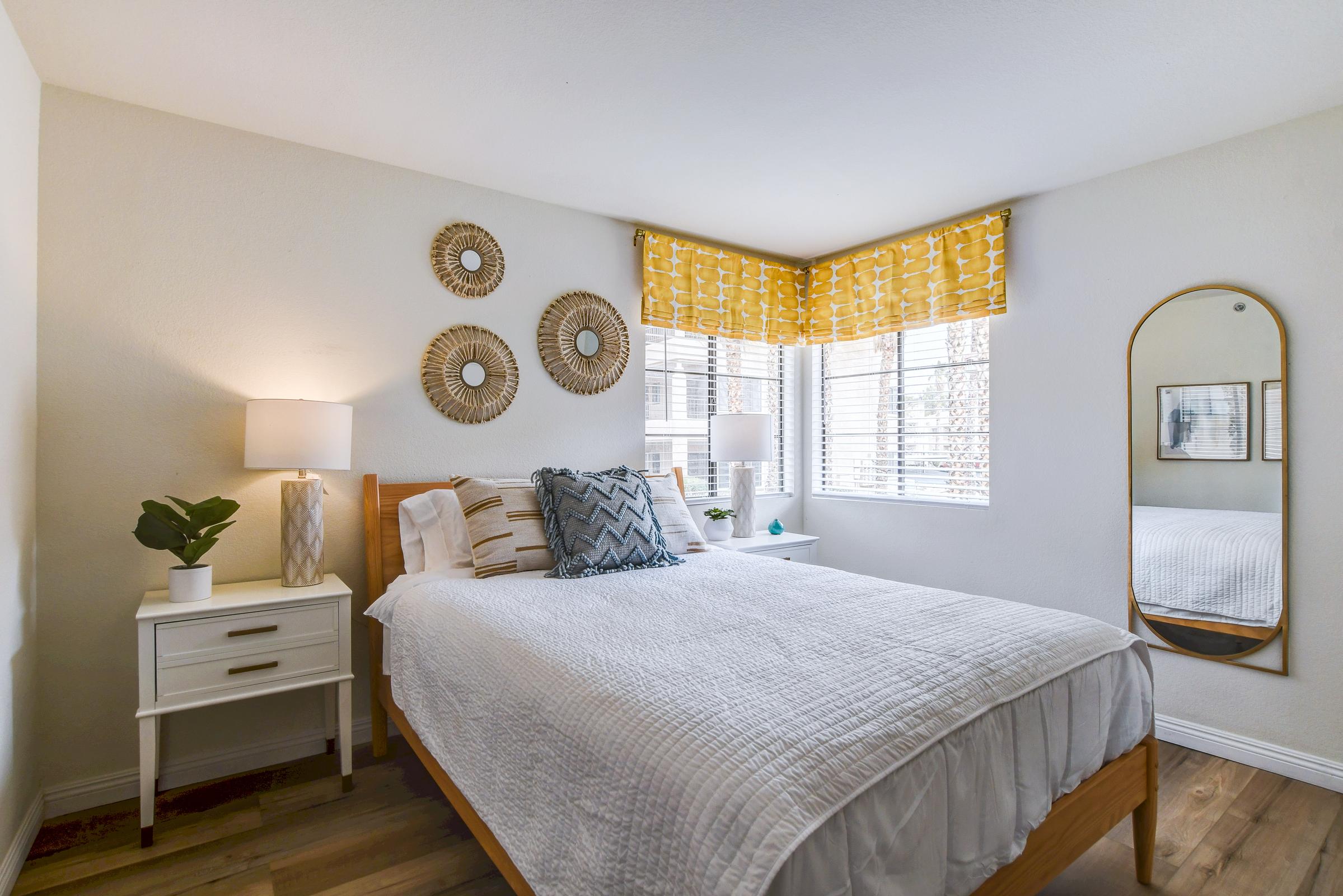 A cozy bedroom featuring a wooden bed with white bedding, two bedside tables with lamps, and decorative wall mirrors. Natural light streams in through window treatments, highlighting a potted plant on the nightstand. The decor is simple yet inviting, creating a warm and tranquil atmosphere.
