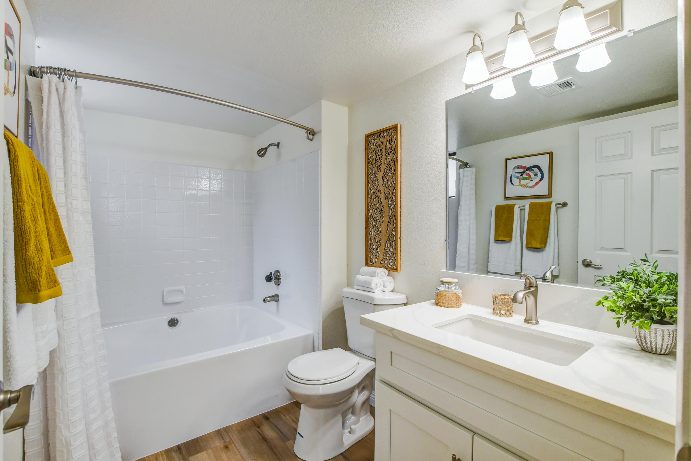A bright, modern bathroom featuring a white bathtub with a shower curtain, a white vanity with a sink and mirror, and decorative elements like towels and a plant. The walls are light-colored and there is wooden flooring, creating a clean and inviting atmosphere.