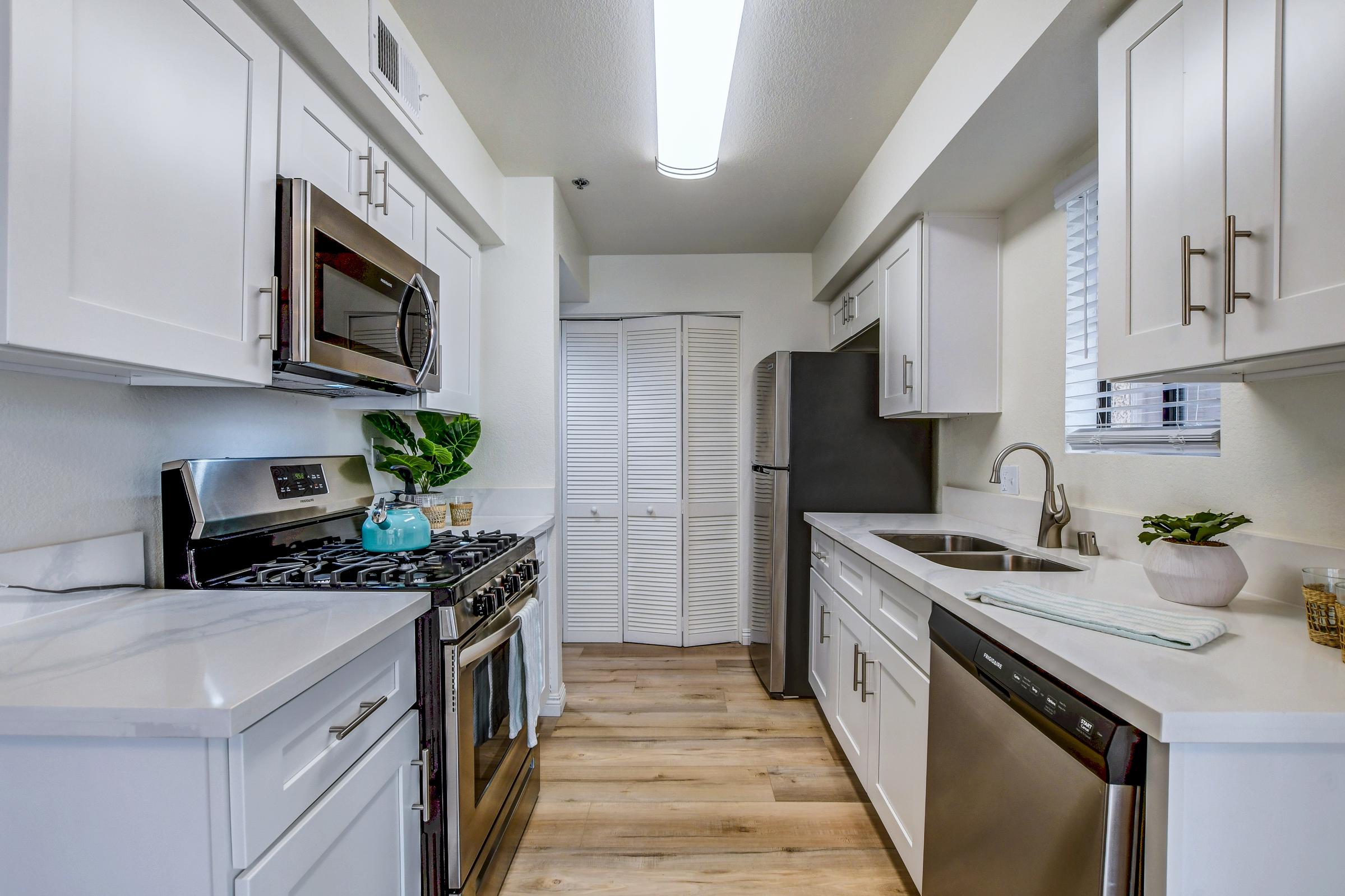 A modern kitchen featuring white cabinetry, stainless steel appliances including a microwave and oven, a gas stove, and a dishwasher. The countertop is made of light-colored material, and there's a sink with a faucet. A teal kettle sits on the stove, and a plant is placed on the counter. Light wood flooring completes the look.