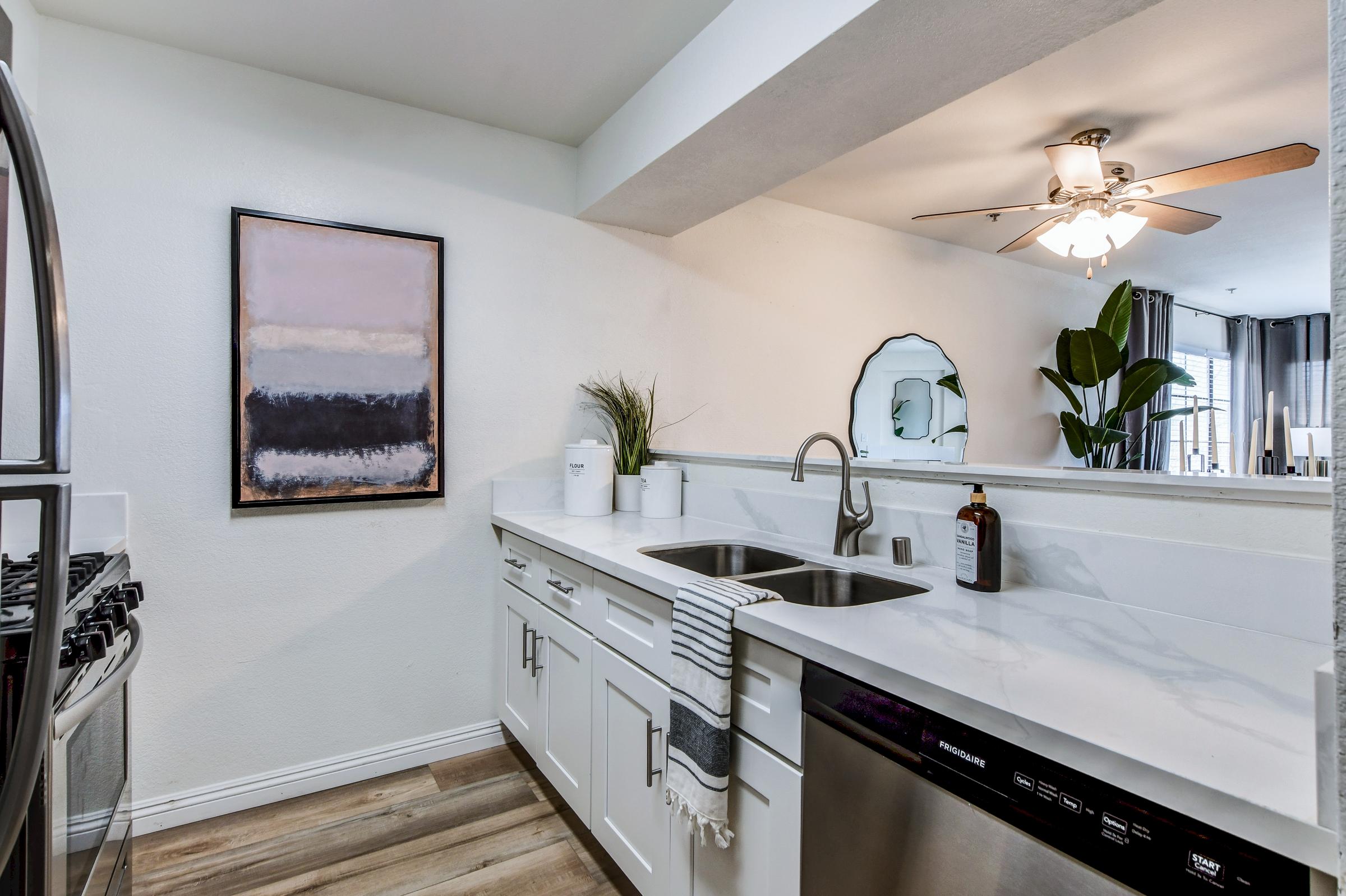 Modern kitchen featuring sleek white cabinetry, stainless steel appliances, and a double sink. A decorative painting hangs on the wall, and a potted plant adds a touch of greenery. The room is well-lit by a ceiling fan and natural light from nearby windows.