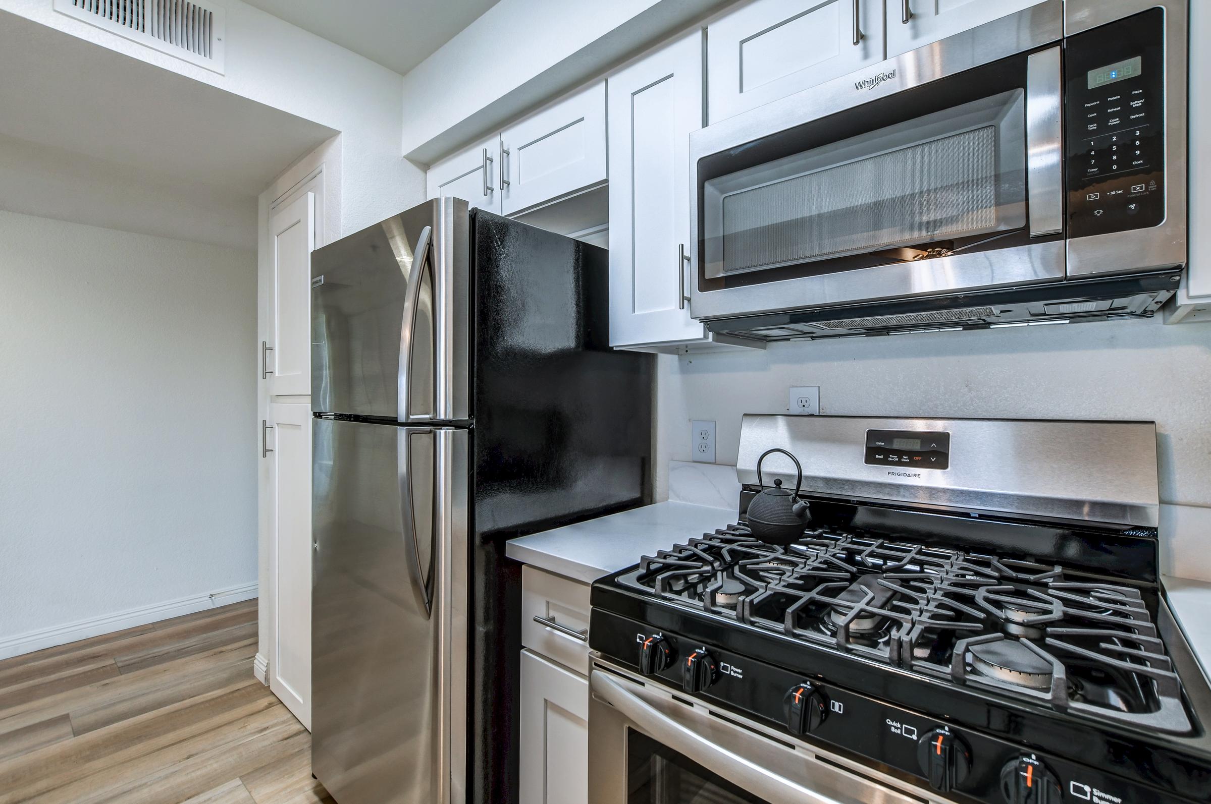 Modern kitchen featuring stainless steel appliances including a gas range, microwave, and refrigerator. The cabinetry is white, with a clean, light color scheme and wooden flooring, creating a bright and inviting atmosphere. The layout is functional and well-organized, ideal for cooking and entertaining.