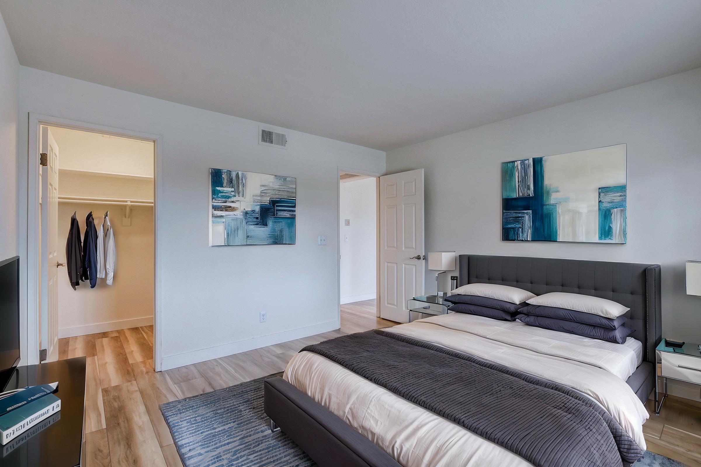 A modern bedroom featuring a neatly made bed with gray and white linens, two abstract paintings on the walls, a nightstand with a lamp, and a flat-screen TV. A closet is visible in the open doorway, and the room has light wood flooring and neutral-colored walls.