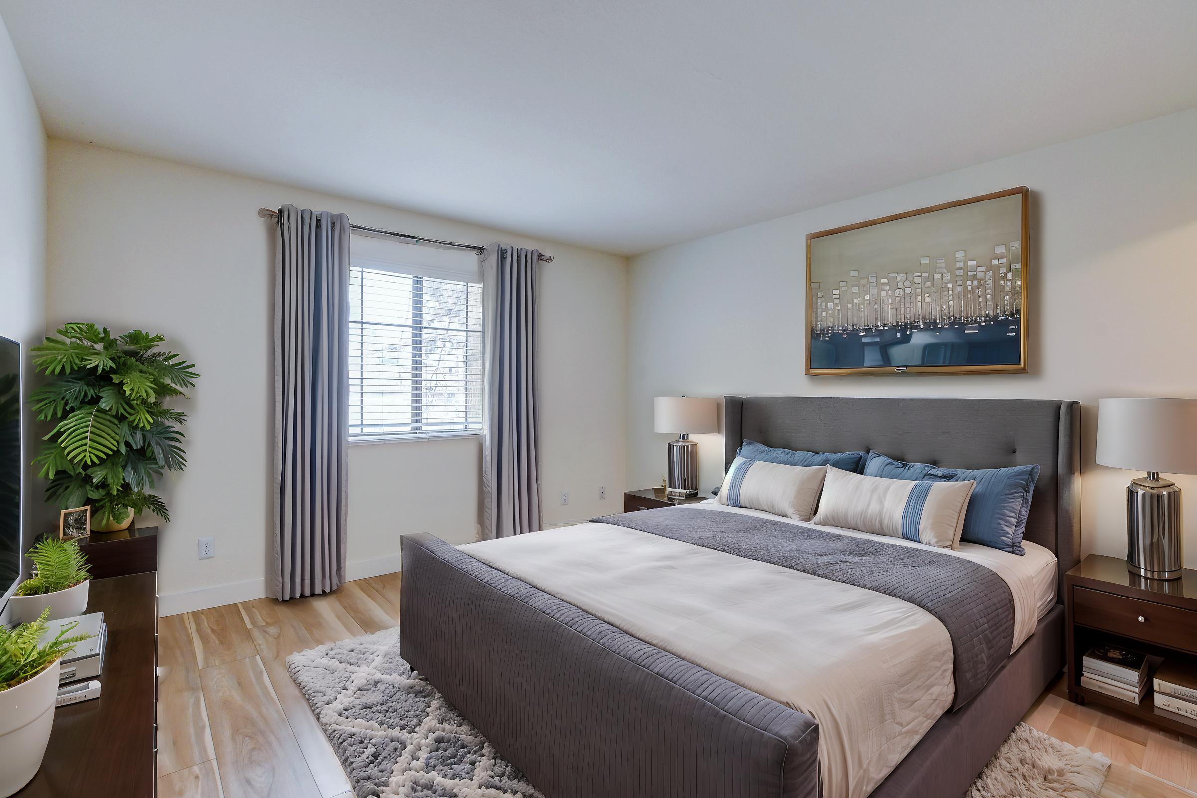 A modern bedroom featuring a queen-sized bed with a gray upholstered headboard, layered with white and gray bedding. There are two bedside tables with lamps, a large framed artwork on the wall, a window with light-colored drapes, and a small plant by the dresser, enhancing the contemporary aesthetic.