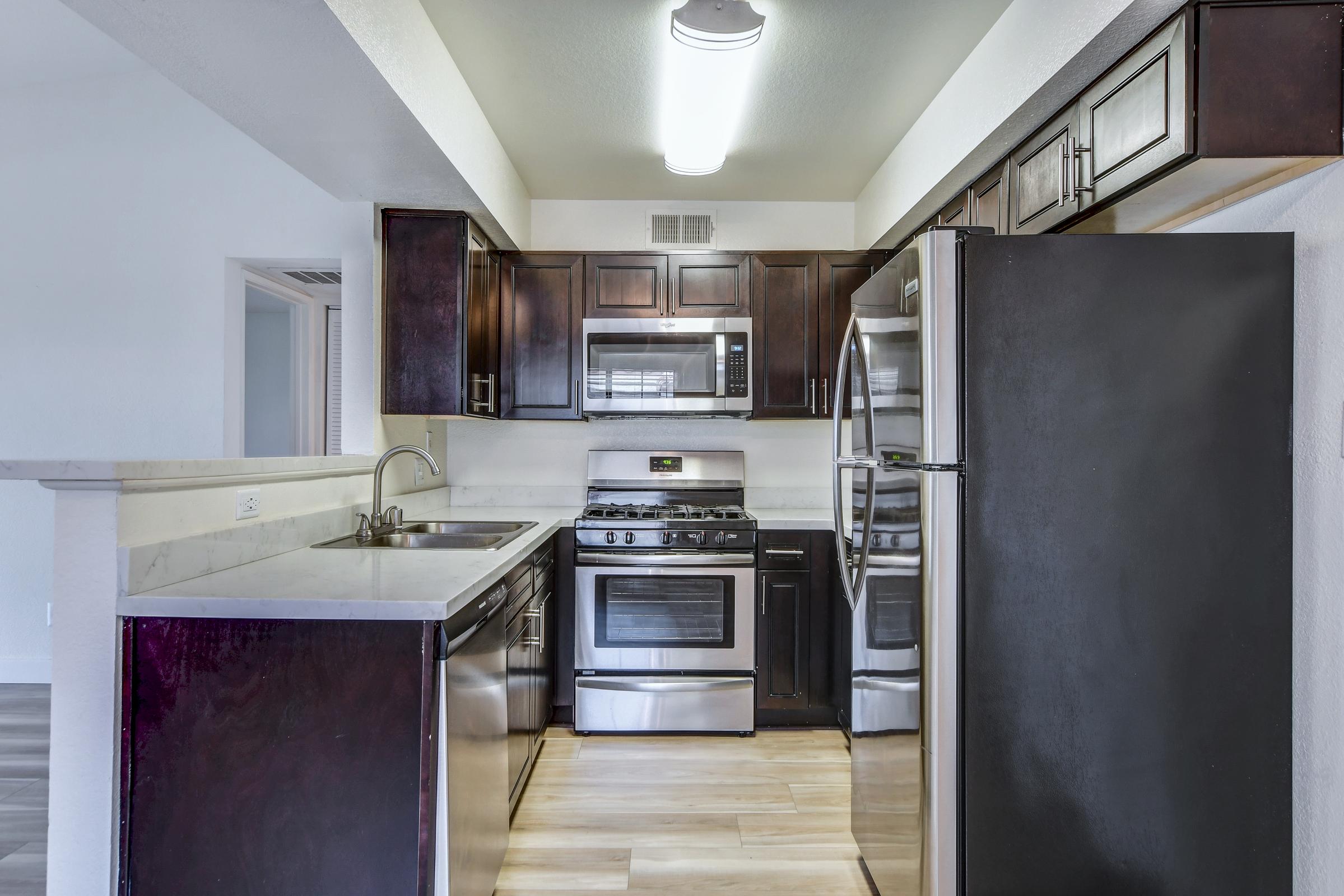 Modern kitchen with dark wood cabinets, stainless steel appliances, and a marble countertop. Features a gas stove, microwave, and refrigerator. The layout is open, with natural light coming from a nearby window. Floor is light-colored wood, creating a contrast with the cabinetry.