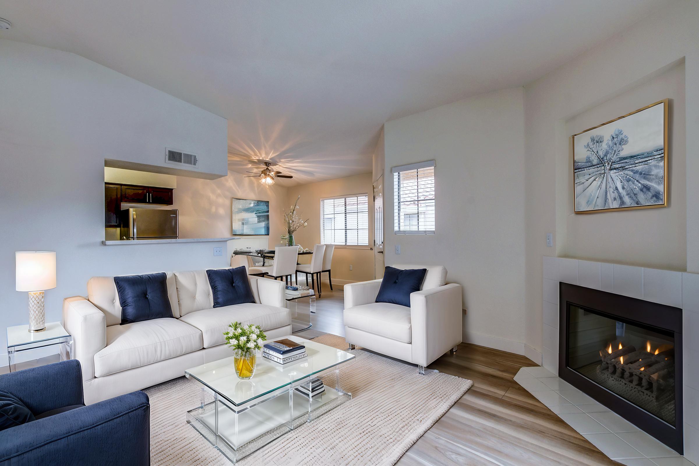A modern living room featuring a light-colored sofa with dark blue accent pillows, a glass coffee table with books and a vase of flowers on it, and a cozy fireplace. The space is bright, with windows allowing natural light in, and includes a dining area visible in the background.