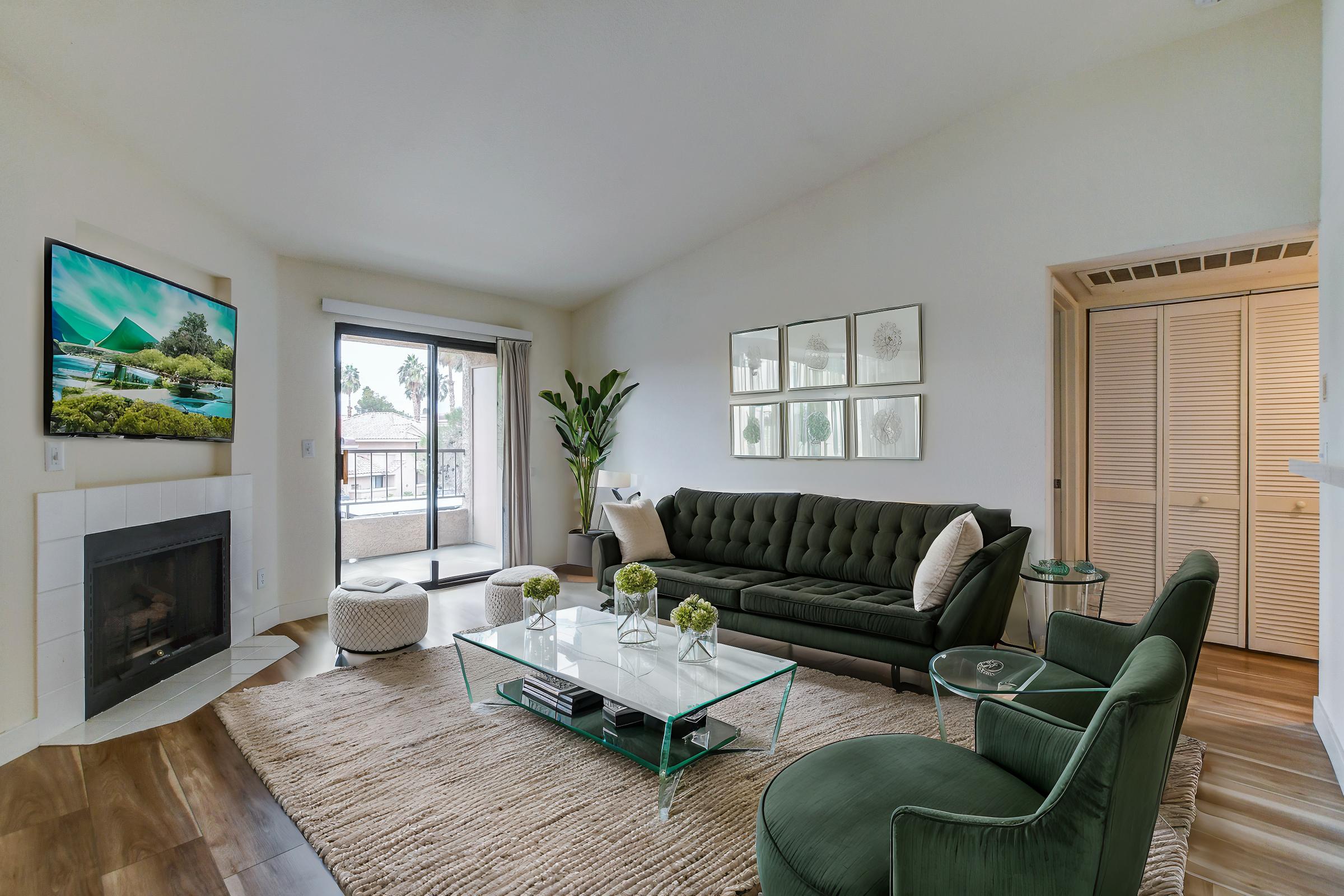 A modern living room featuring a green tufted sofa and a glass coffee table. There's a large flat-screen TV mounted on the wall, a potted plant in the corner, and a sliding glass door that opens to a balcony. The space has a cozy, bright ambiance with wood flooring and neutral decor.