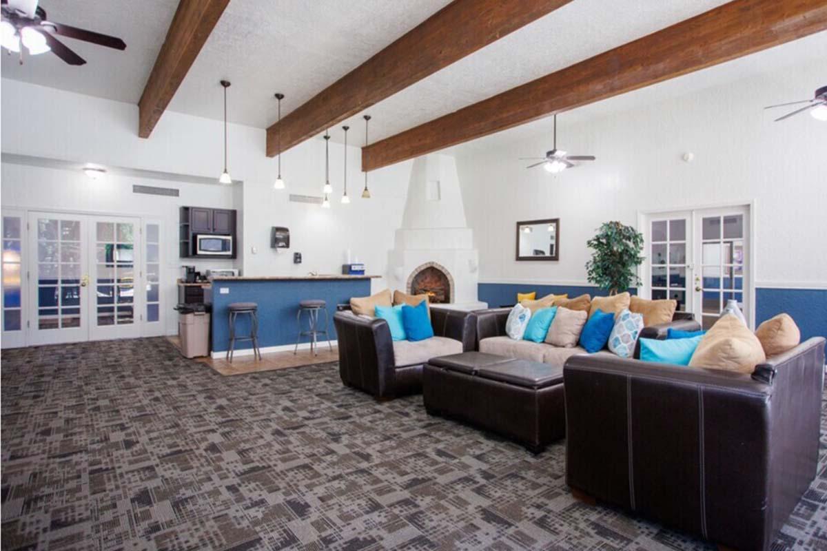 A spacious living area featuring a cozy configuration of dark leather couches and a large coffee table with colorful throw pillows. The room has high ceilings with wooden beams, a fireplace, and a kitchenette area visible in the background. Well-lit with ceiling fans and modern décor, it offers an inviting atmosphere.