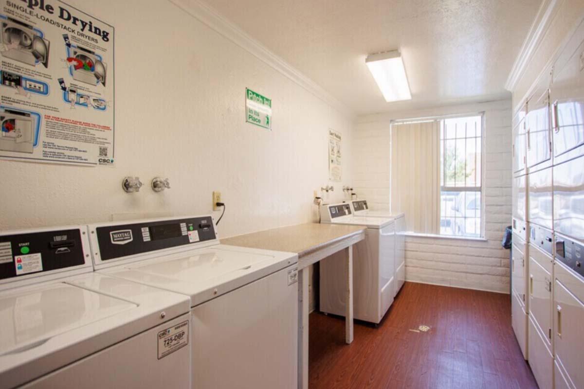 A clean and well-lit laundry room featuring multiple washing machines and dryers along one wall. There are counters for folding clothes and posters on the walls detailing laundry instructions. A window lets in natural light, and the flooring is a light wood finish.