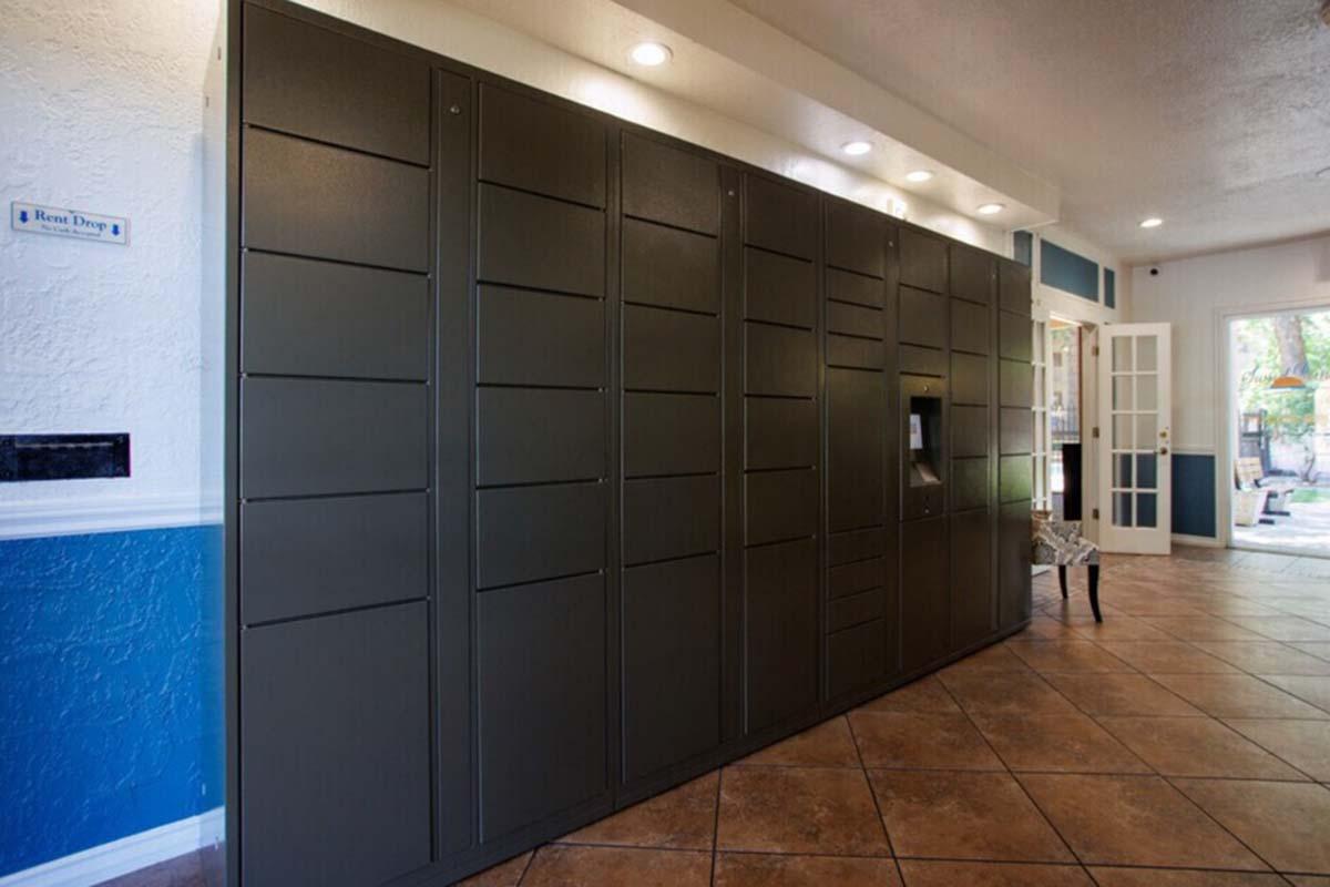 A row of grey delivery lockers installed in a modern interior space. The lockers are neatly aligned against a wall and feature multiple compartments for package delivery. Soft lighting above the lockers enhances the ambiance of the area, which includes a tiled floor and a glimpse of an open door leading outside.