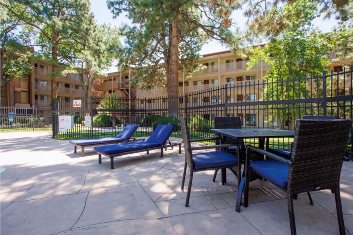 A poolside area featuring two blue lounge chairs and a dining table with blue cushions. In the background, a fenced-in pool area is surrounded by greenery and tall trees, with apartment buildings visible. The setting is bright and inviting.