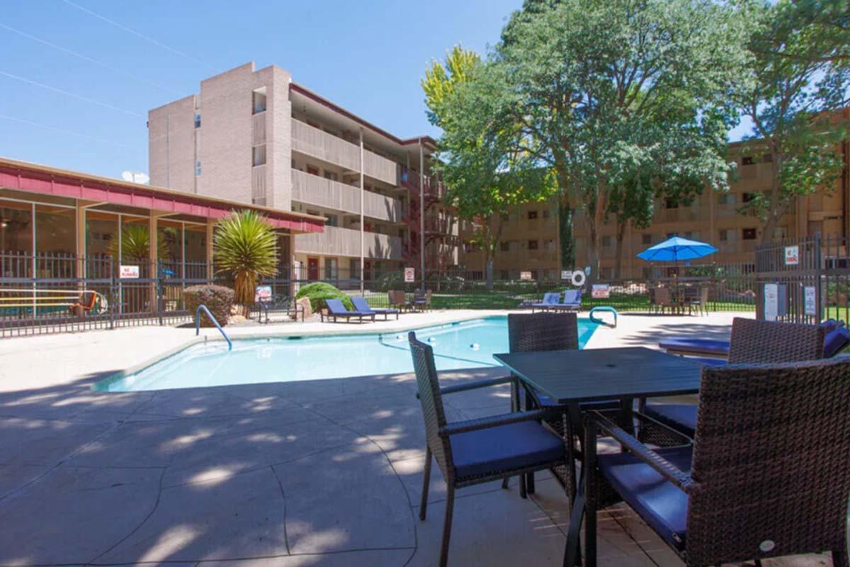 A clear blue swimming pool surrounded by lounge chairs and tables, with a few palm trees in the background. In the distance, a multi-story brick building is visible, and sun umbrellas provide shaded areas. The scene is bright and inviting, showcasing a relaxing outdoor space.