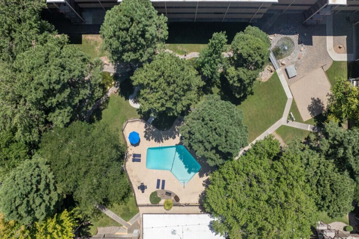 An aerial view of a landscaped area featuring a rectangular swimming pool surrounded by green trees. There are several lounge chairs near the pool, and a blue umbrella provides shade. Pathways and grassy spaces are visible, along with an area paved with stones.