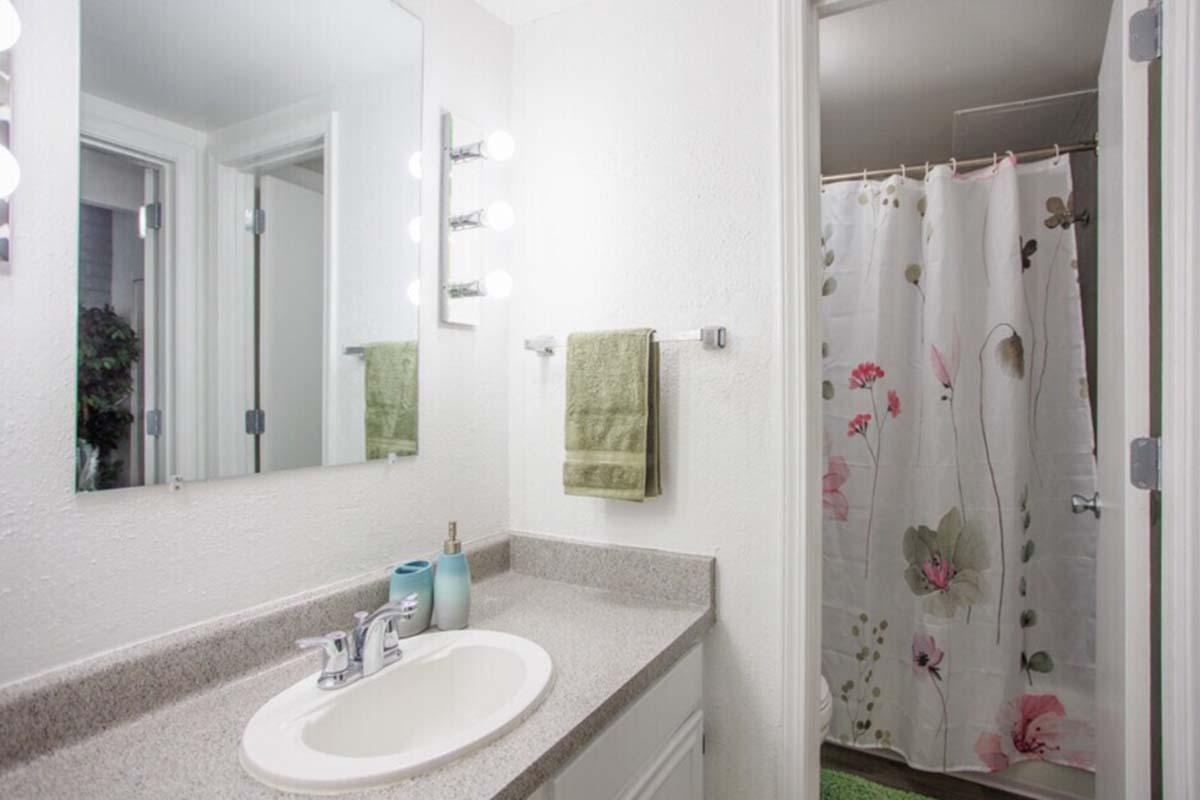 A modern bathroom featuring a gray countertop with a sink and a mirror above it. There's a light fixture with multiple bulbs, a green towel hanging on a rack, and a shower area with a floral curtain. The walls are painted white, and there's a small green mat on the floor.