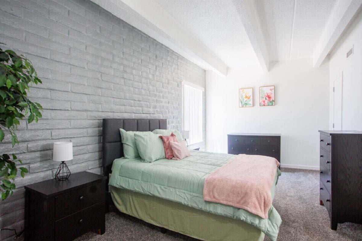 A modern bedroom featuring a gray brick accent wall, a queen-sized bed with a mint green bedspread and pink throw blanket, and two decorative pillows. There are two dark wooden nightstands with lamps on either side of the bed. The room has light-colored carpet and two framed floral artworks on the wall.