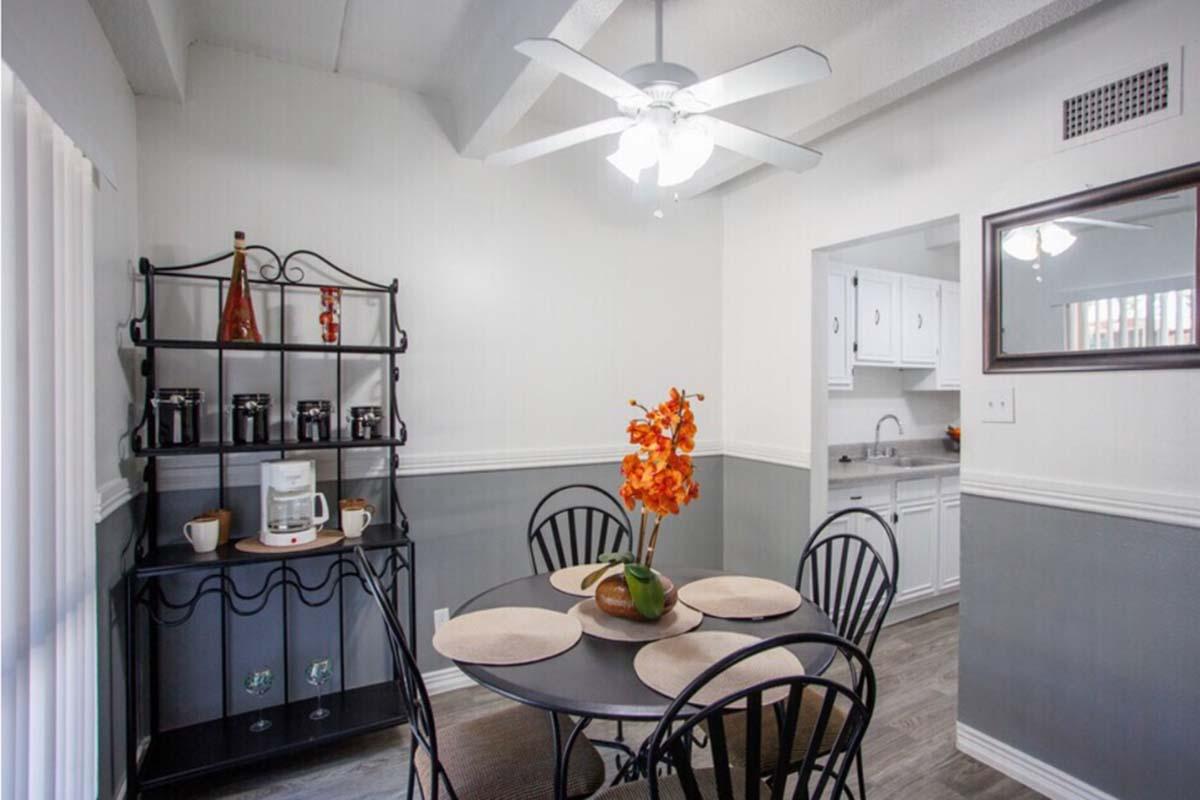 A cozy dining area featuring a round black table with four chairs, set with placemats. A small decorative shelf holds jars and a coffee maker. A vase with orange flowers sits on the table. The walls are painted light gray with white trim, and a ceiling fan is visible above. The adjoining kitchen can be seen in the background.