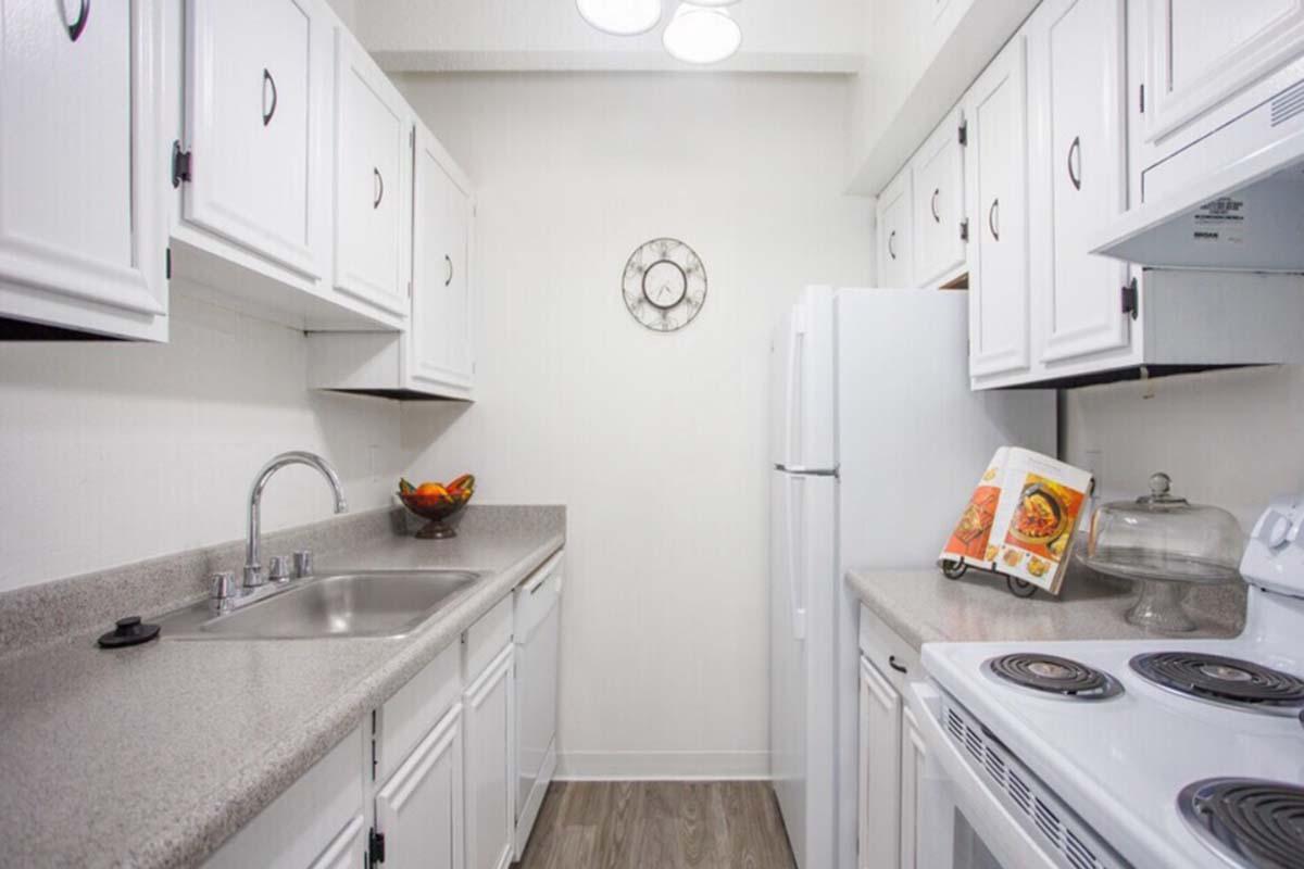 A bright, clean kitchen featuring white cabinetry, a double sink, and a countertop. The space includes a refrigerator, an oven with a stovetop, and a wall-mounted clock. A bowl of fruit is placed on the counter, and cooking magazines are propped up for easy reference. The flooring is a light wood finish.