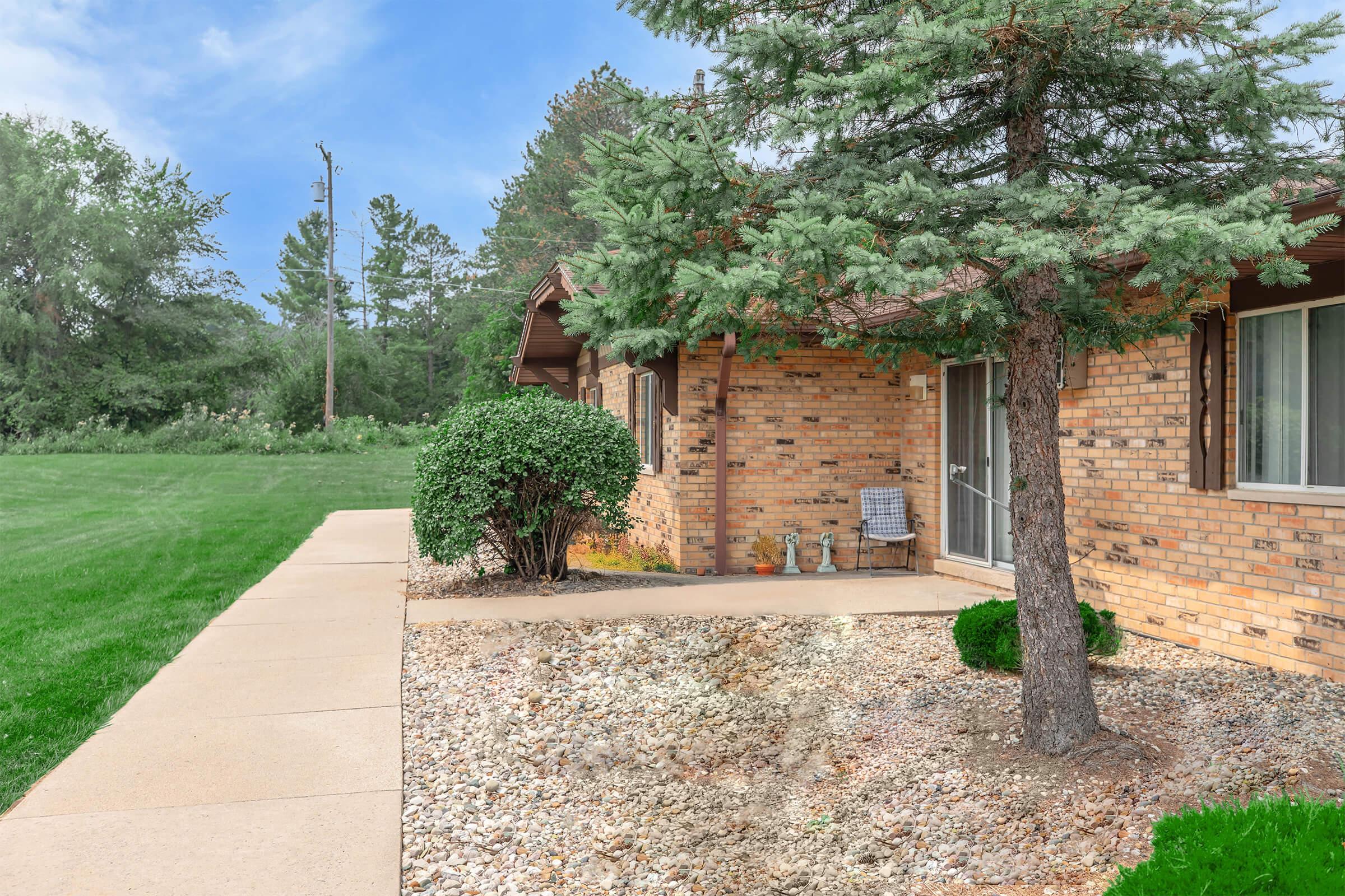 Side view of a brick house with a concrete pathway leading to the entrance. A lush green lawn is visible on the left, with a few shrubs and a small tree in the foreground. The surrounding area features trees and a grassy field in the background under a clear blue sky.