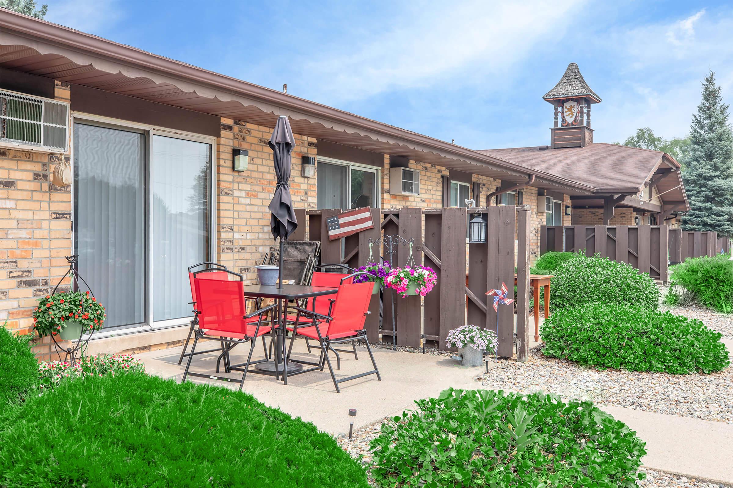 A cozy outdoor patio with a small round table and four red chairs. The area is surrounded by lush green shrubs and flowers, featuring a decorative fence. In the background, a building with a clock tower is visible, alongside air conditioning units and large windows.