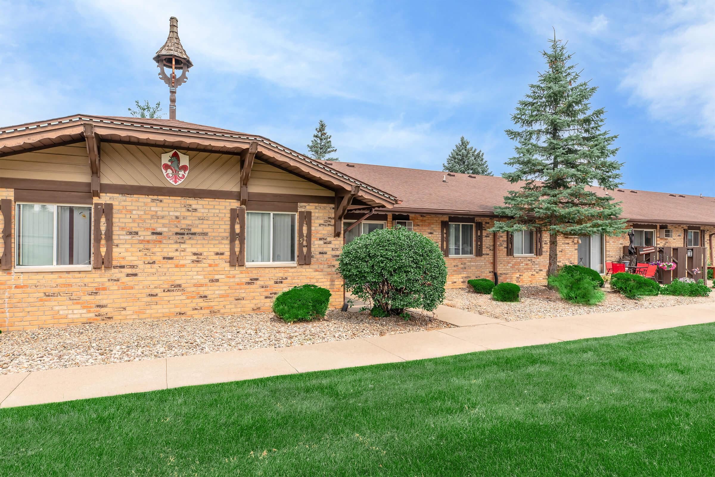 A brick building with a pitched roof, featuring a decorative cupola and a shield emblem above a front entrance. The landscaped area includes neatly trimmed bushes and grass, with another building visible in the background. The sky is clear and blue, suggesting a pleasant day.